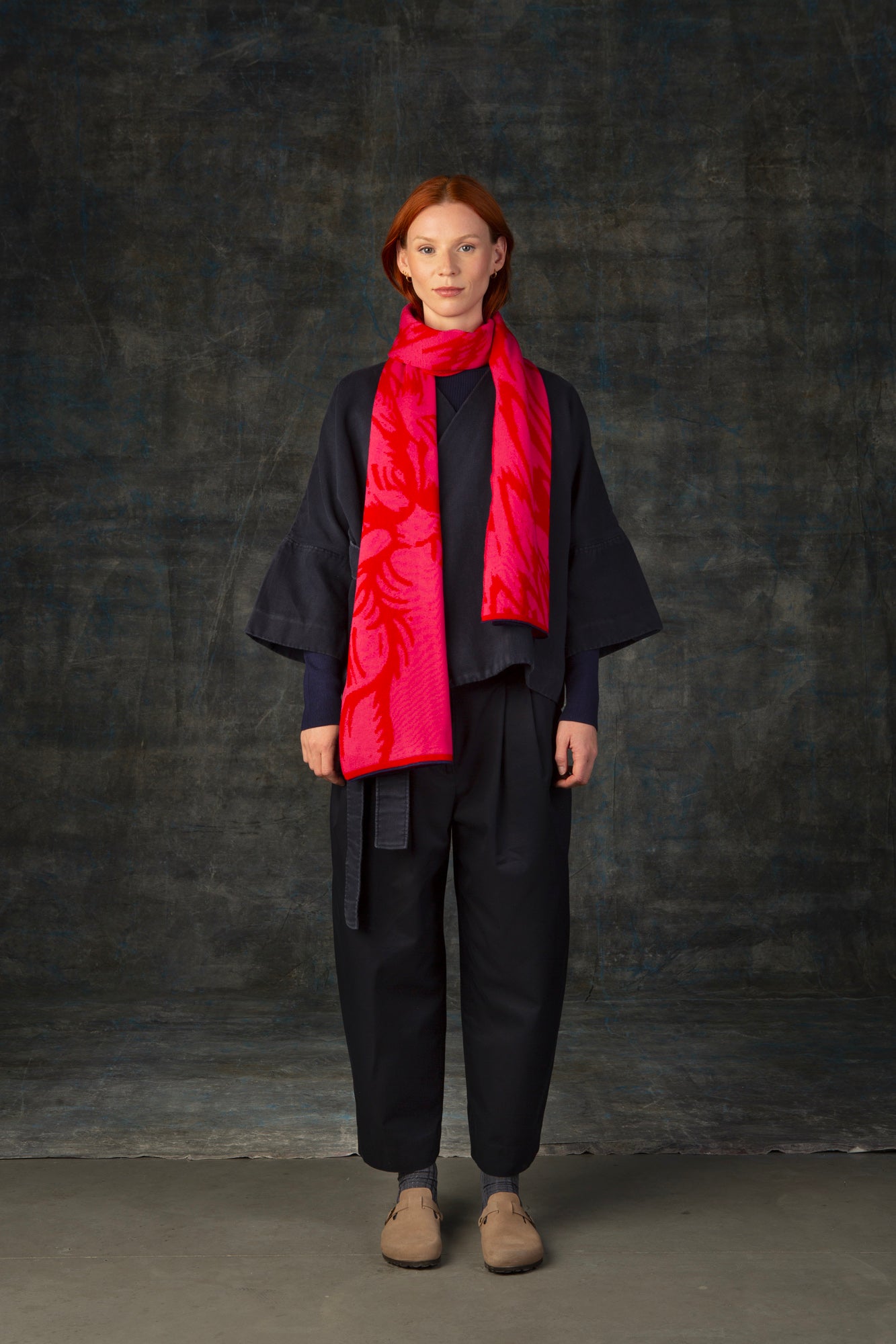 Female model wearing a dark blue outfit with a bright red and pink maxi scarf with nature-inspired pattern draped around her neck, against a dark background.