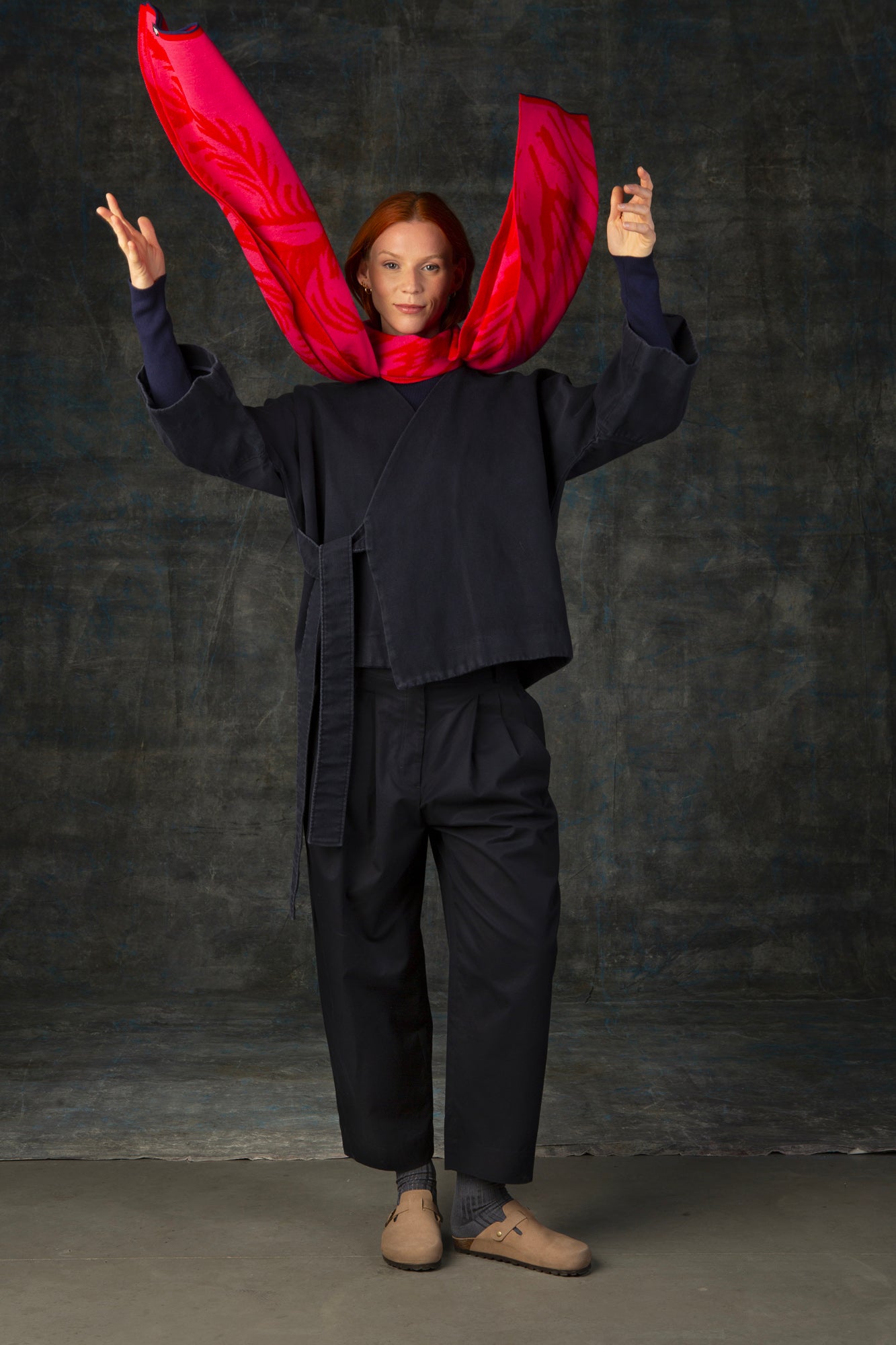 Female model wearing a dark blue outfit throwing up the ends of a bright red and pink maxi scarf, with nature-inspired pattern.
