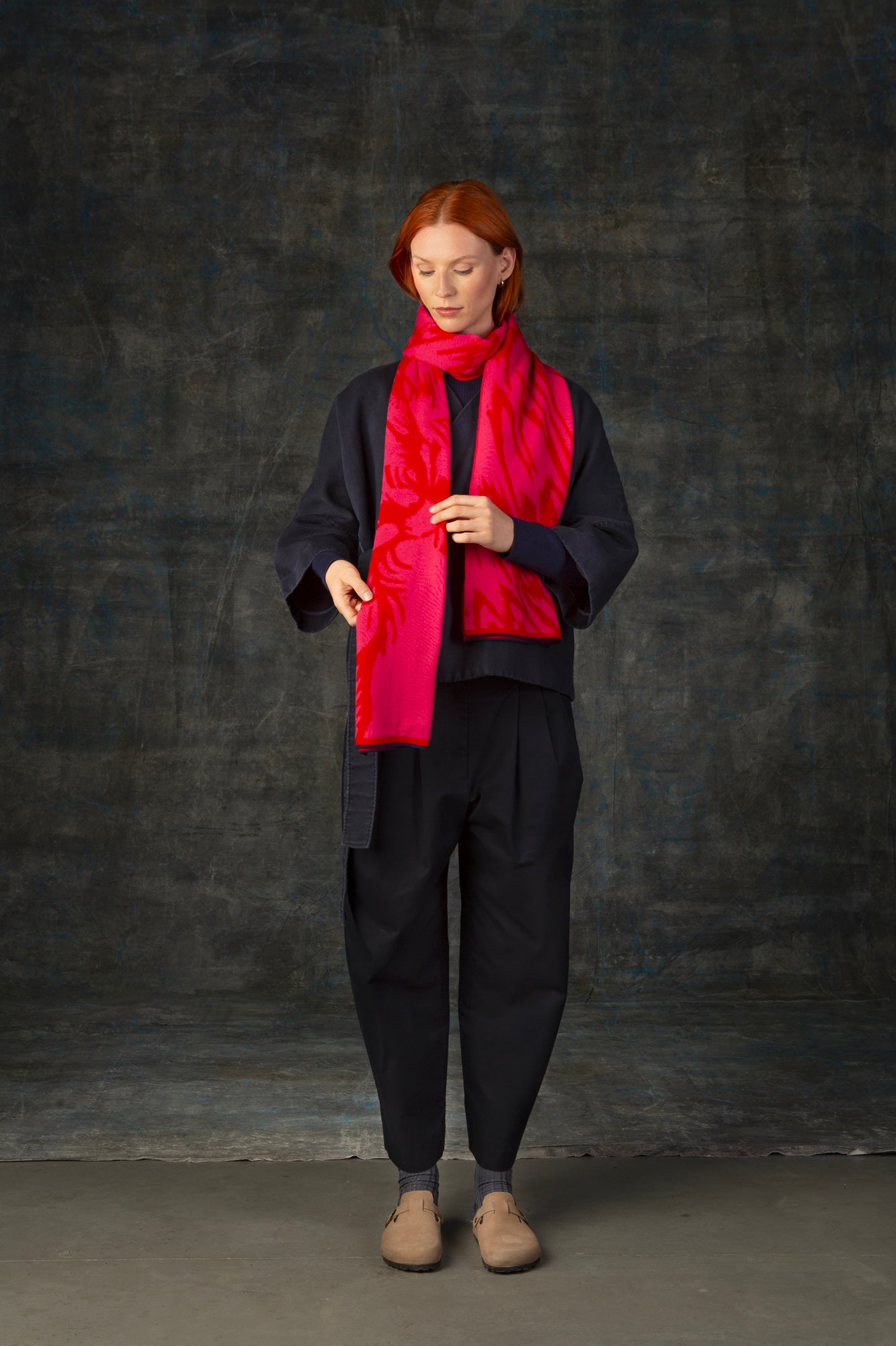 Female model wearing a dark blue outfit with a bright red and pink maxi scarf with nature-inspired pattern draped around her neck, against a dark background.