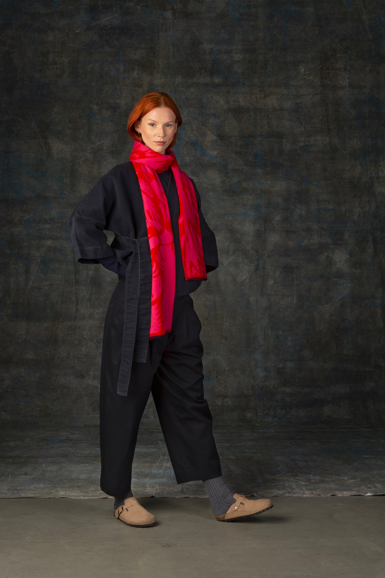 Female model wearing a dark blue outfit with a bright red and pink maxi scarf with nature-inspired pattern draped around her neck, against a dark background.