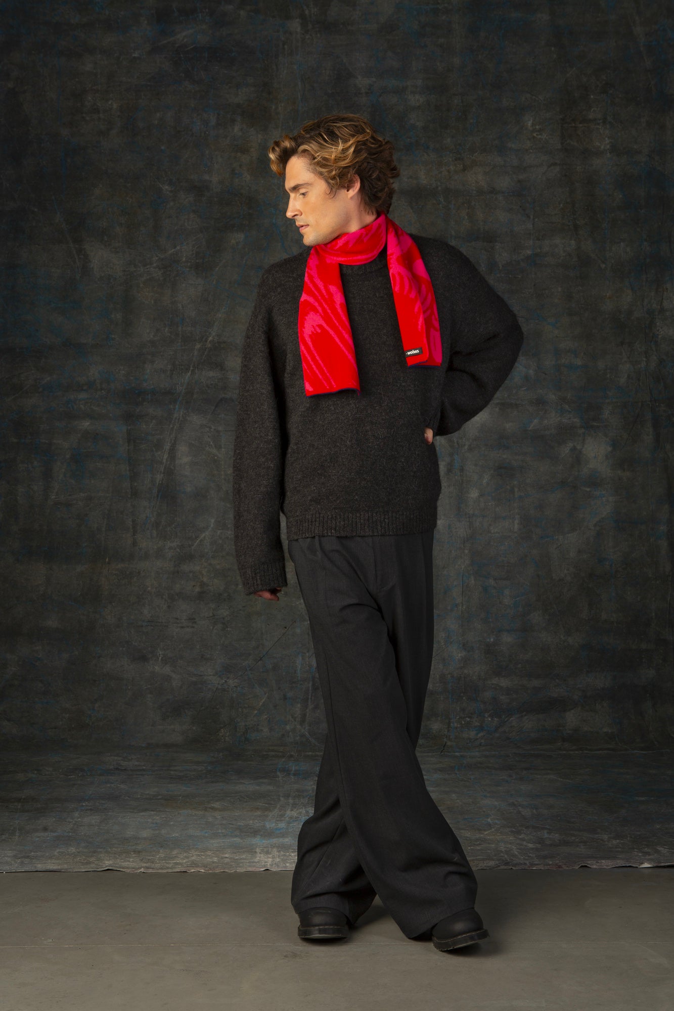 Male model wearing a dark blue outfit with a bright red and pink mini scarf, with nature-inspired pattern, draped around his neck, against a dark background.