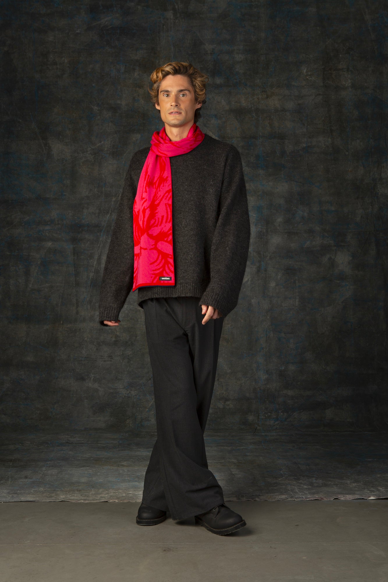 Male model wearing a dark grey outfit with a bright red and pink midi scarf, with nature-inspired pattern draped over his shoulder, against a dark background.