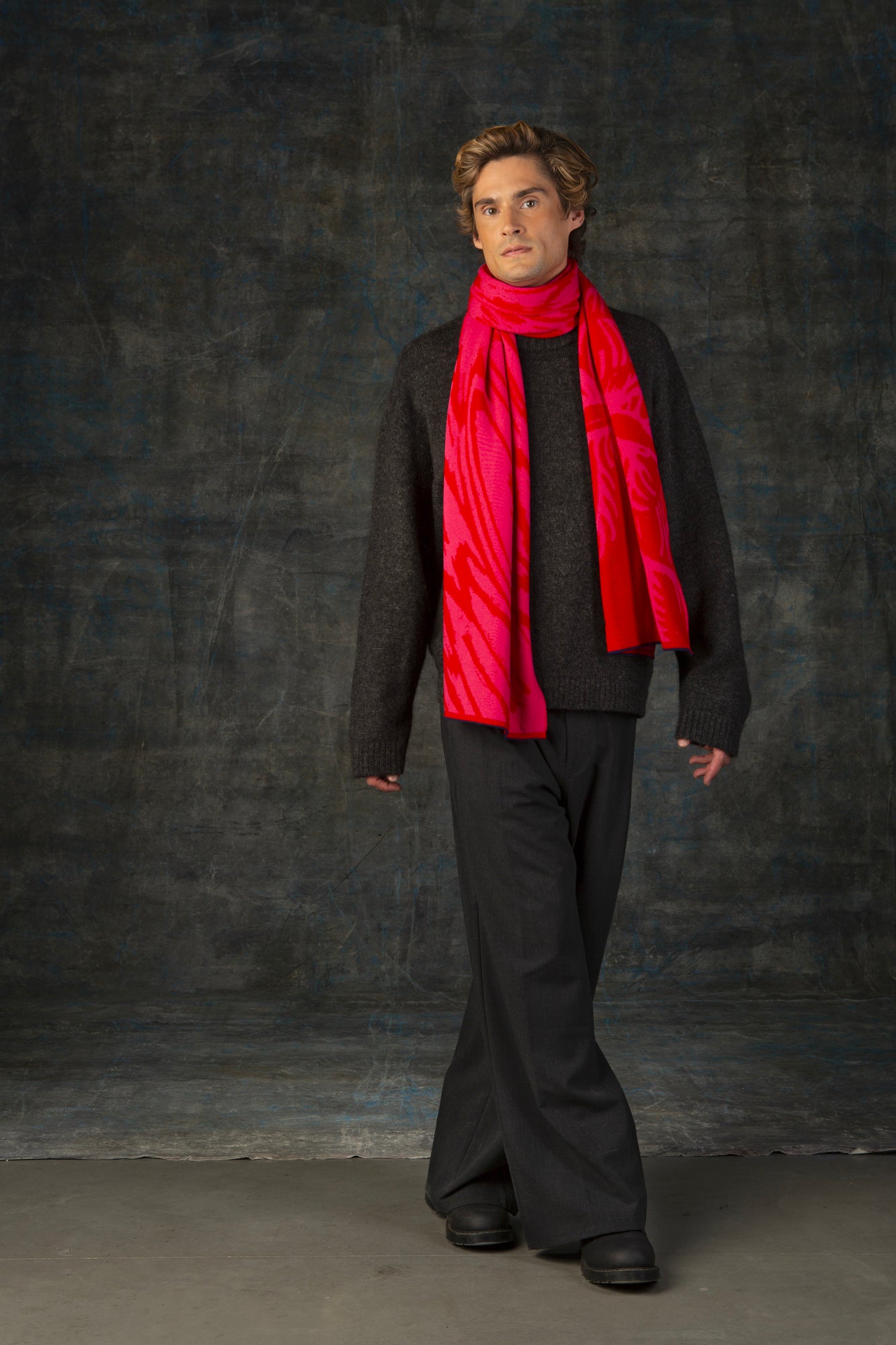 Male model wearing a dark grey outfit with a bright red and pink maxi scarf with nature-inspired pattern draped around his neck, against a dark background.