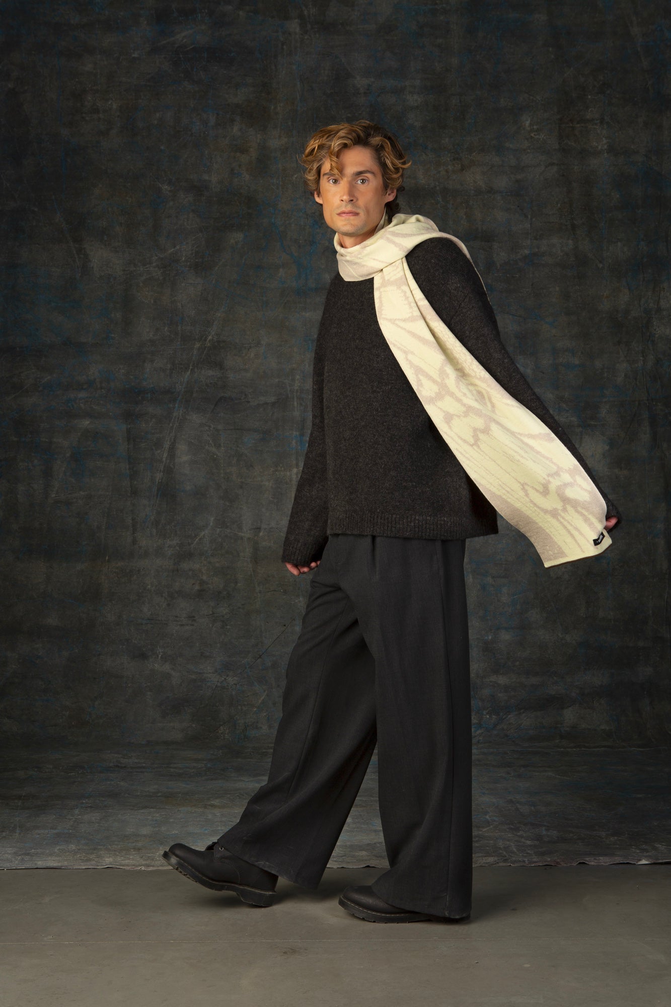 Male model wearing a grey outfit with an ecru midi scarf draped over his shoulder, against a dark background.