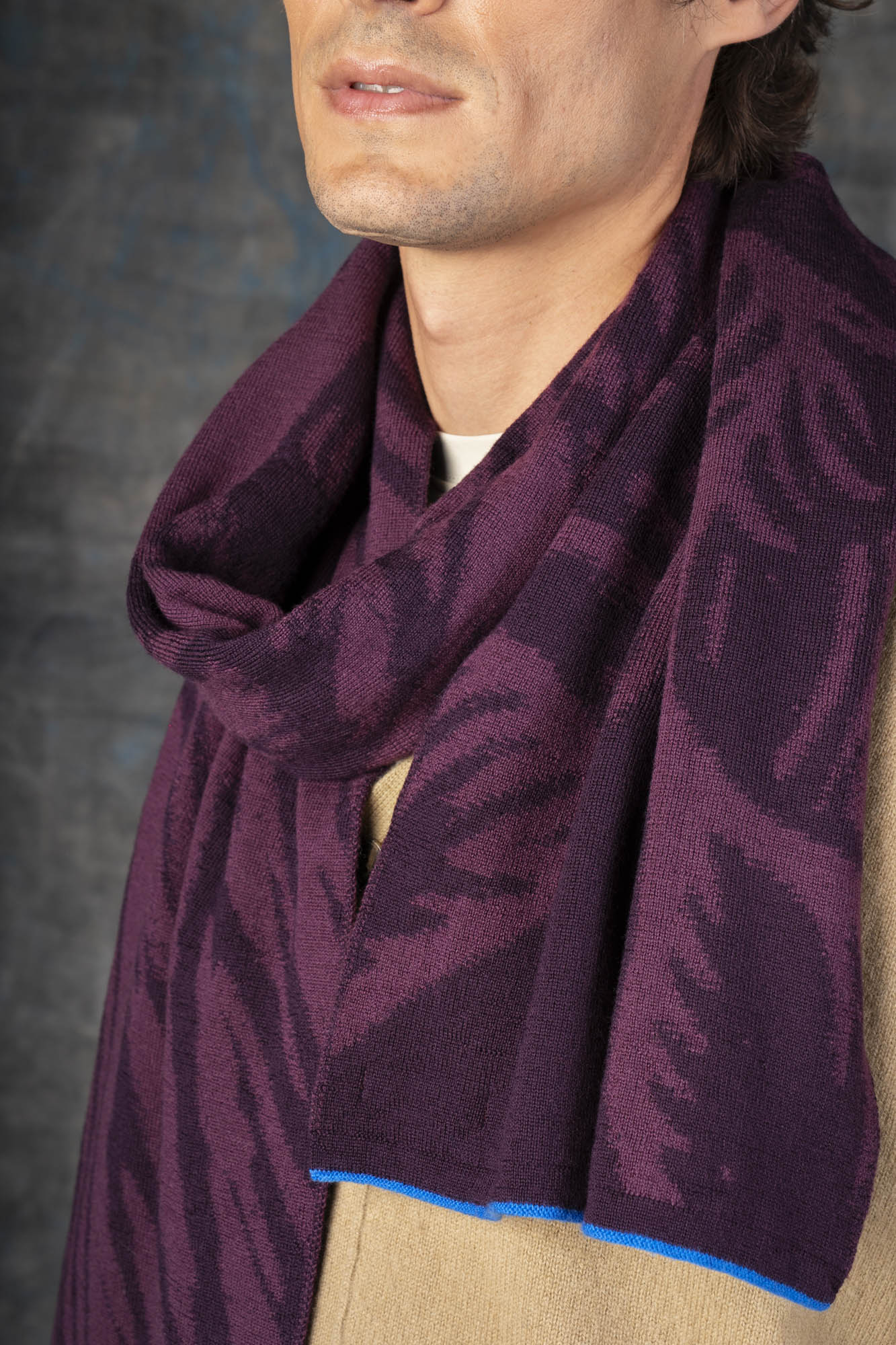 Close-up of a male model wearing a light brown outfit with a dark purple midi scarf, with nature-inspired pattern, loosely draped around his neck.