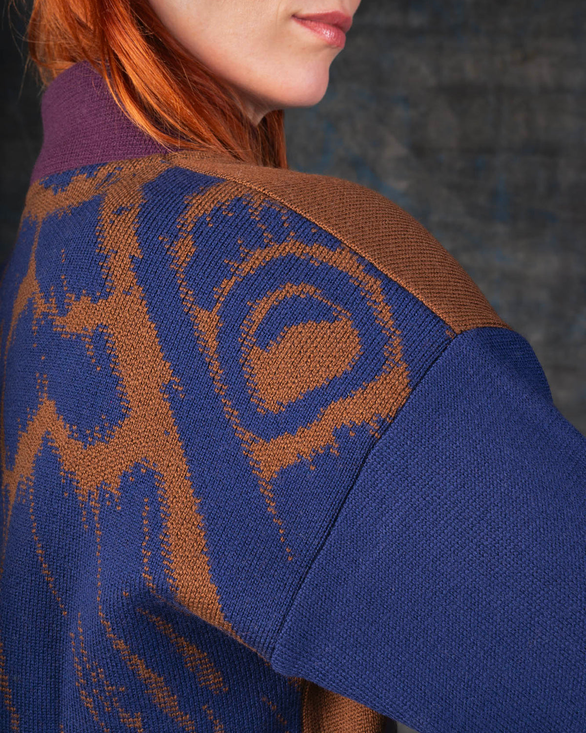 Close-up of a person wearing a brown bomber jacket with dark blue sleeves and purple collar. There is an organic pattern inspired by a butterfly's wings on the back of the jacket.