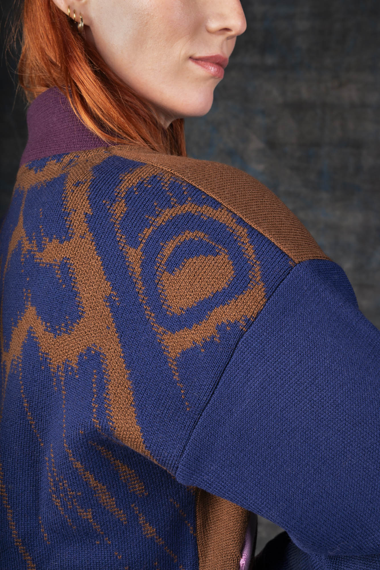 Close-up of a person wearing a brown bomber jacket with dark blue sleeves and purple collar. There is an organic pattern inspired by a butterfly's wings on the back of the jacket.