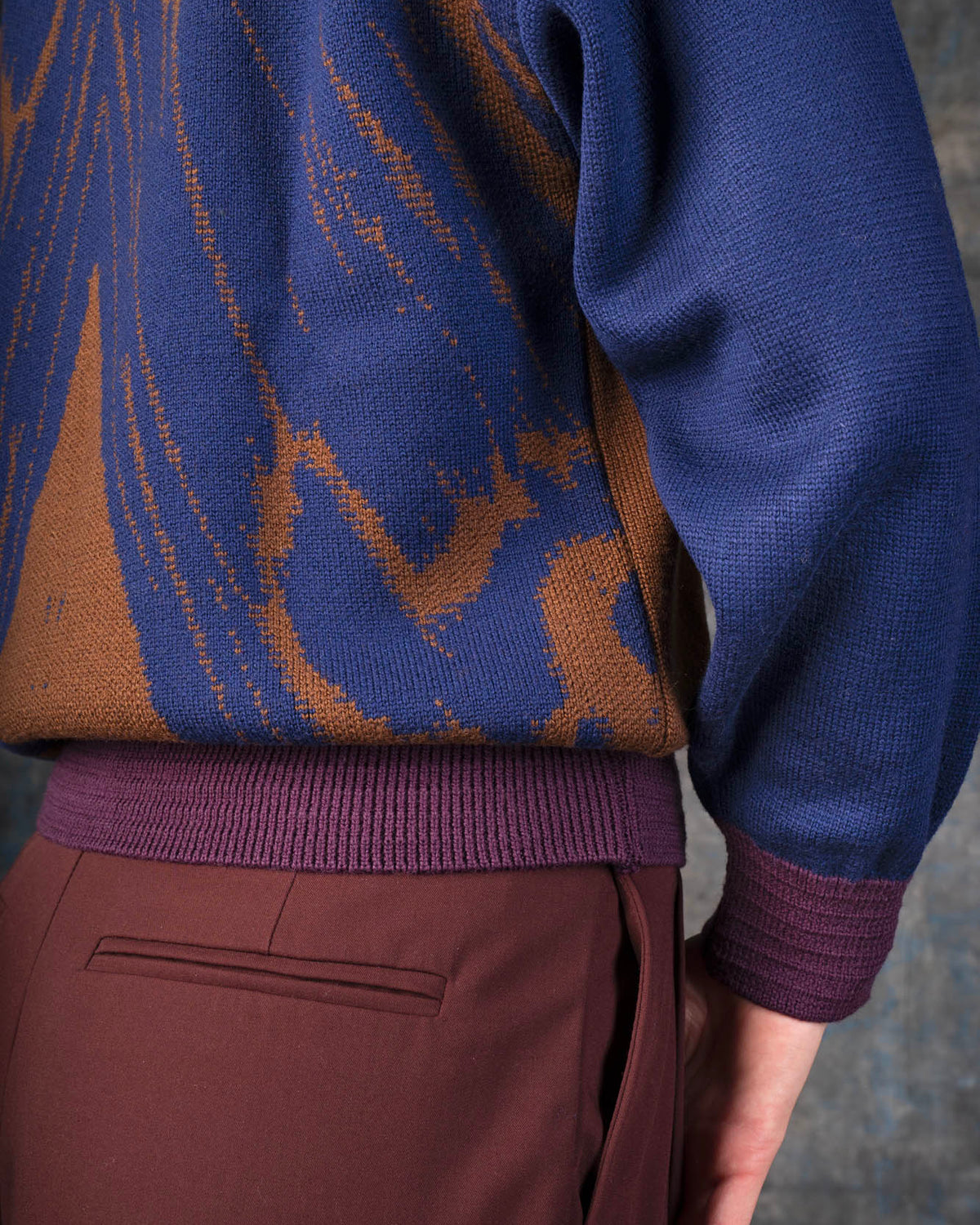 Close-up of a person wearing a brown bomber jacket with dark blue sleeves and purple cuffs. There is an organic pattern inspired by a butterfly's wings on the back of the jacket.