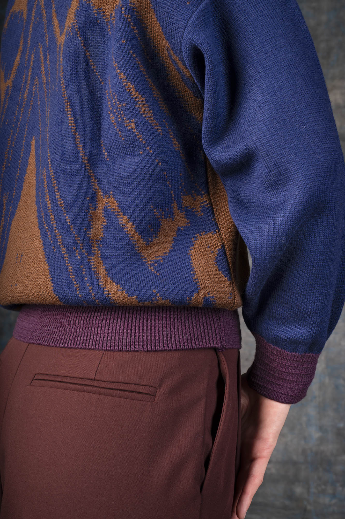 Close-up of a person wearing a brown bomber jacket with dark blue sleeves and purple cuffs. There is an organic pattern inspired by a butterfly's wings on the back of the jacket.