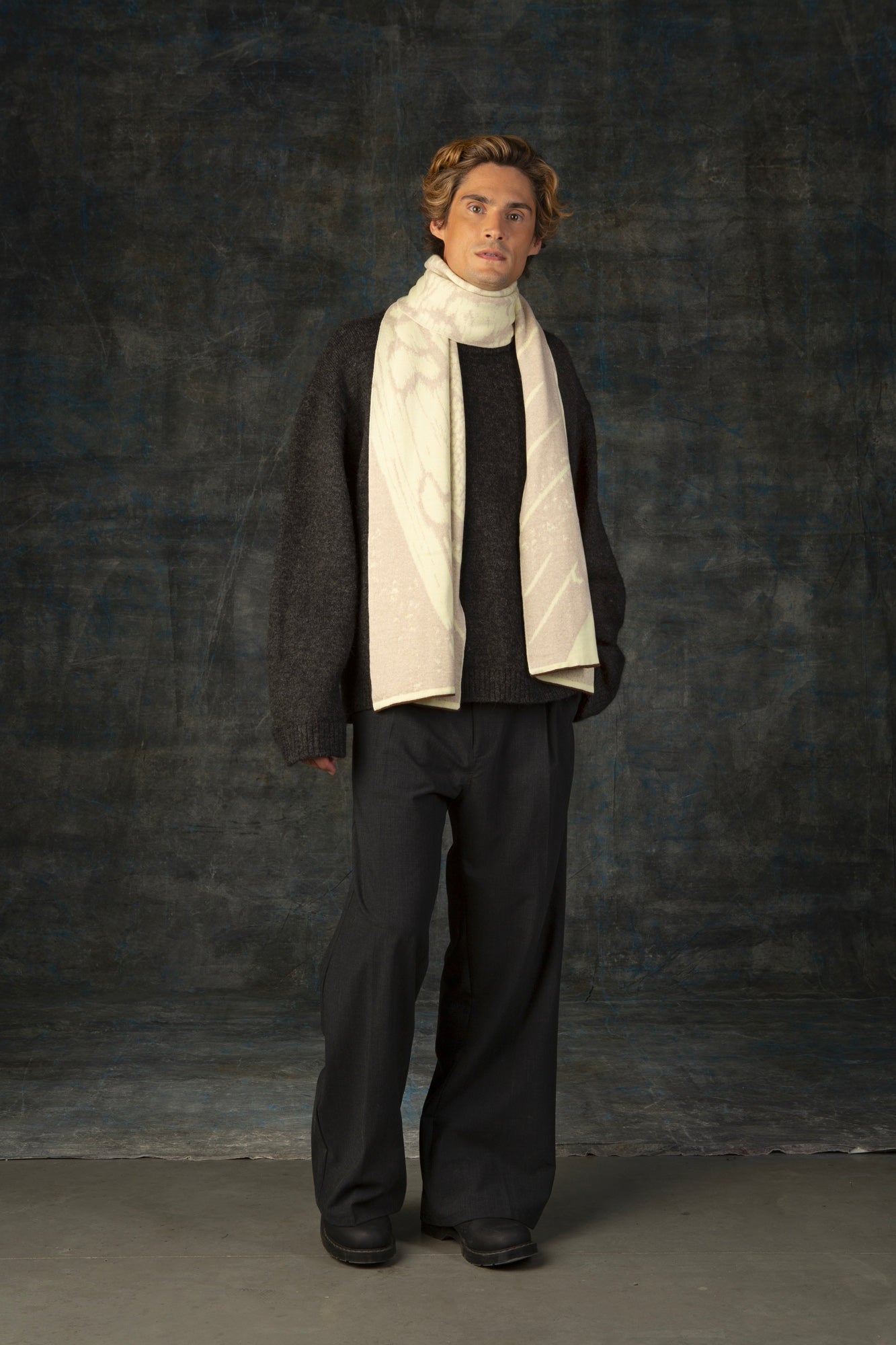 Male model wearing a grey outfit with an ecru maxi scarf draped around his neck, against a dark background.