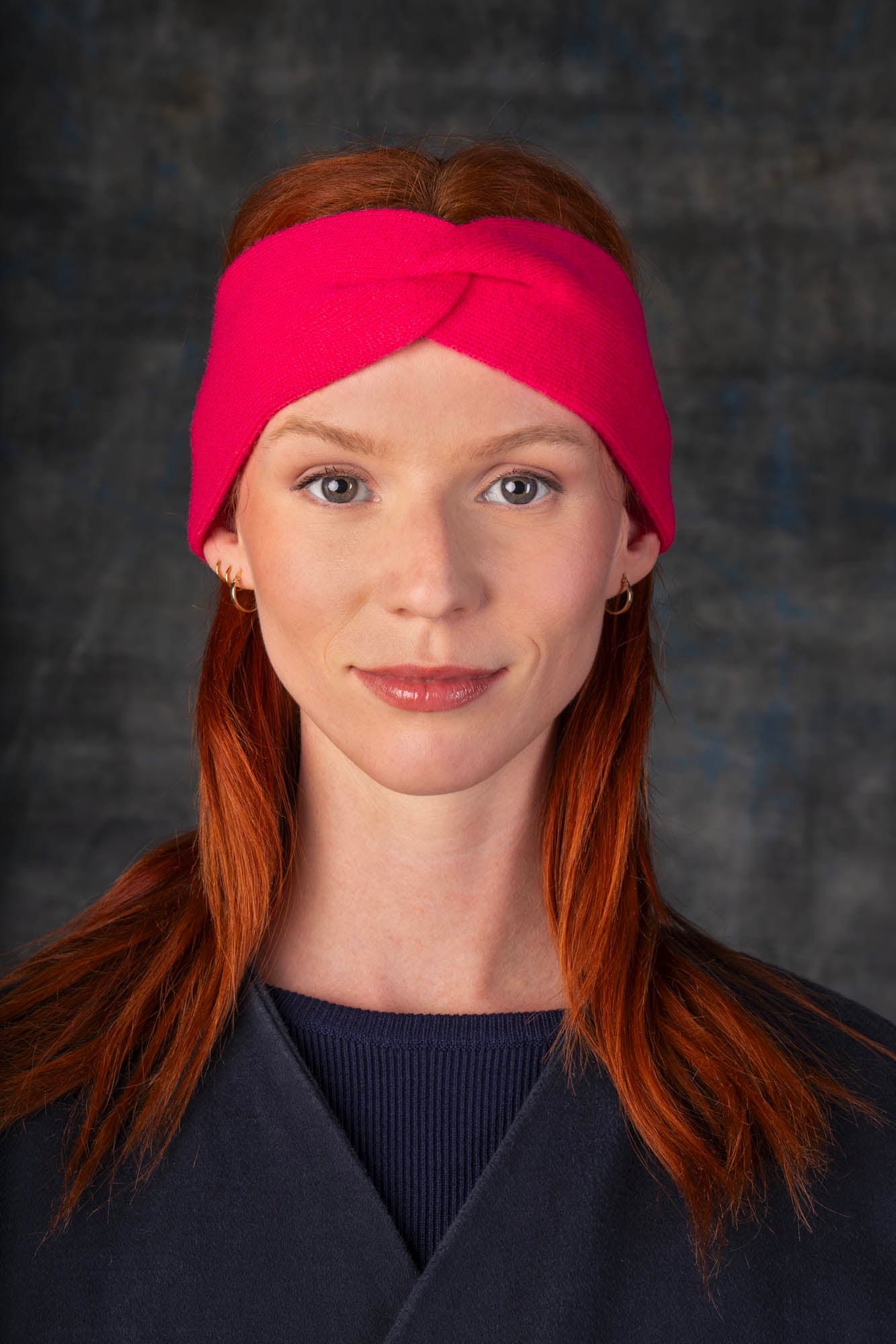 Person wearing a bright pink knit headband and dark blue coat against a dark background.