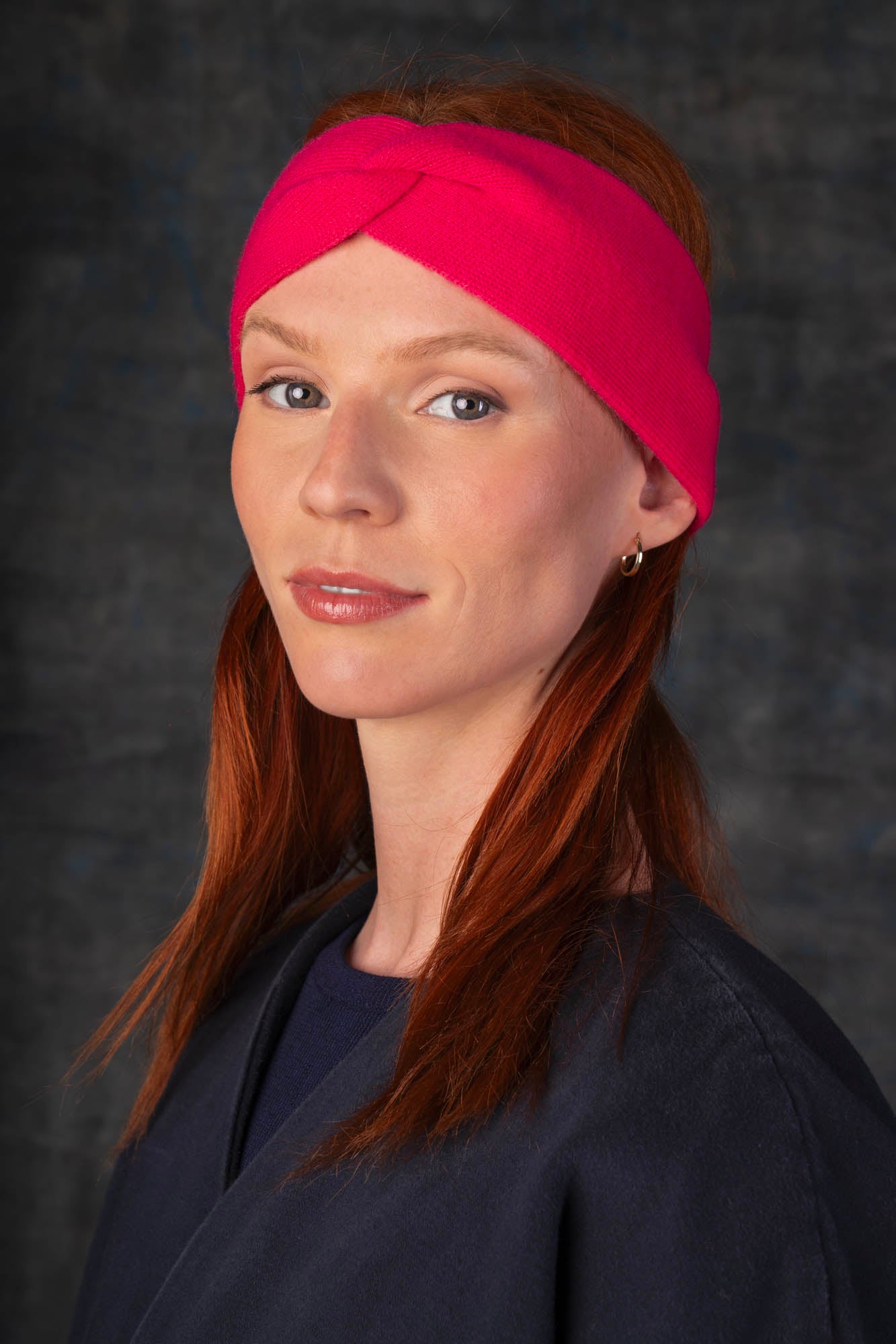 Person wearing a bright pink knit headband and dark blue coat against a dark background.