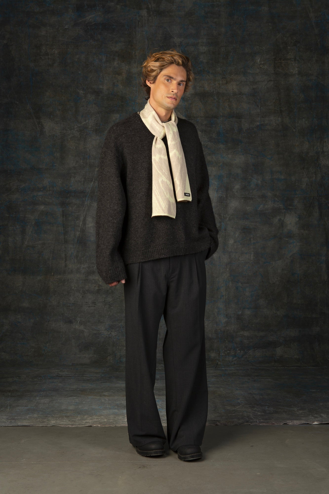 Male model wearing a grey outfit with an ecru mini scarf loosley tied around his neck, against a dark background.