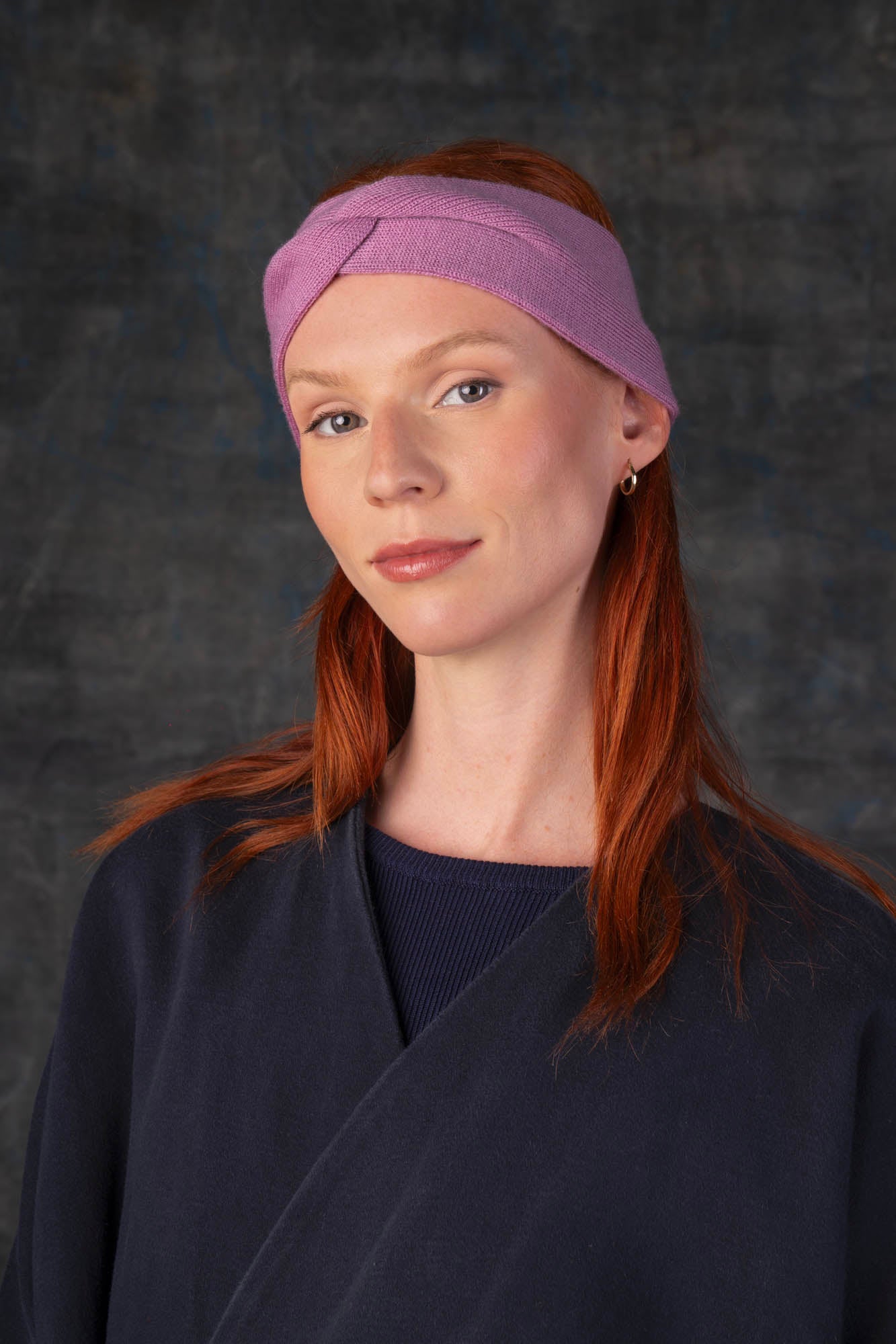 Person wearing a mauve pink knit headband and dark blue coat against a dark background.