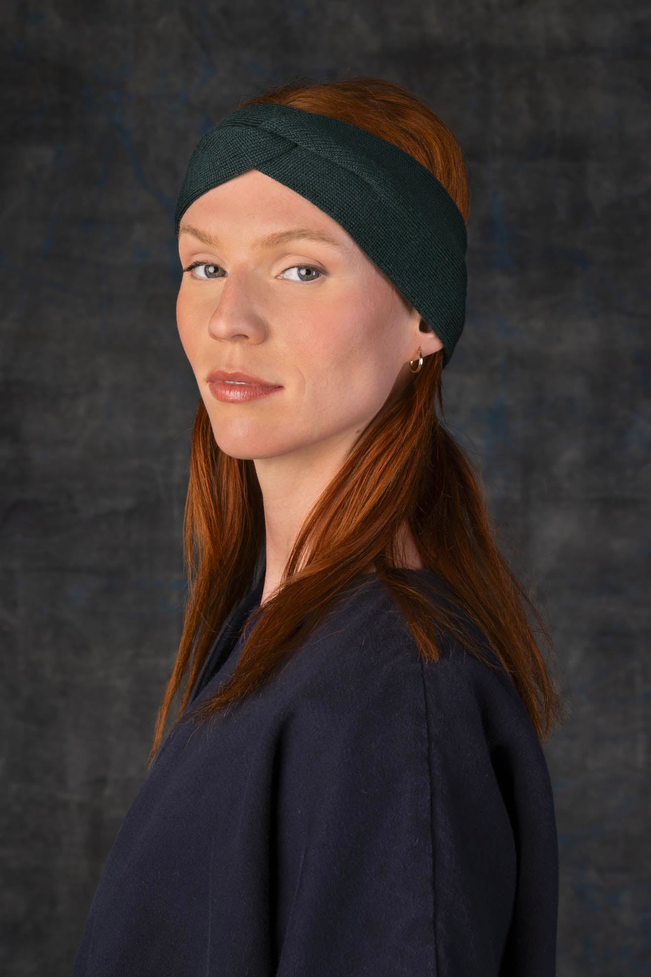 Person wearing a dark green knit headband and dark blue coat against a dark background.