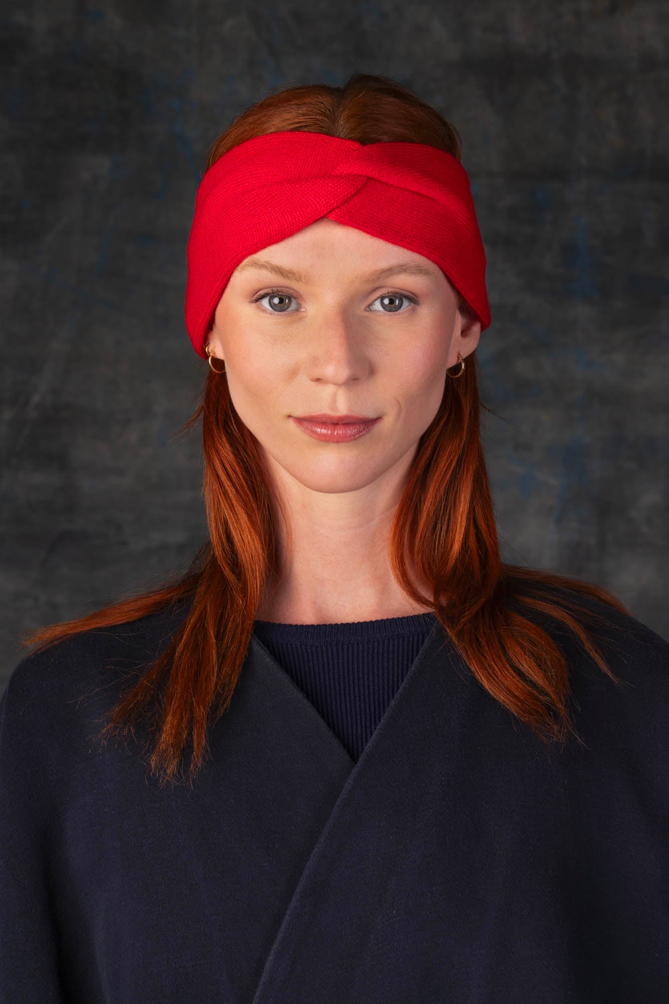 Person wearing a bright red knit headband and dark blue coat against a dark background.