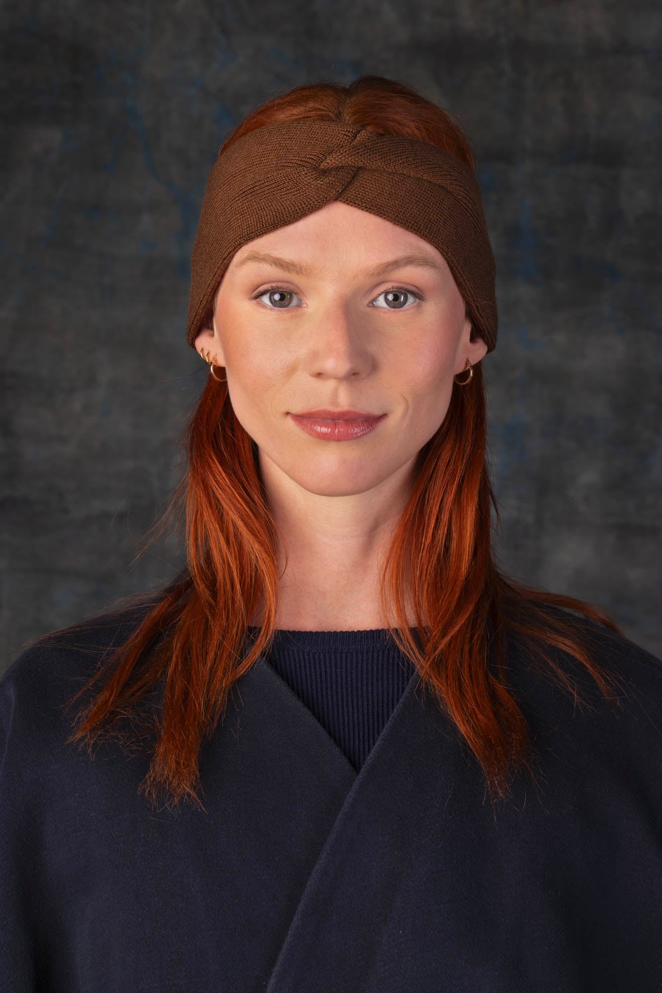 Person wearing a cacao brown knit headband and dark blue coat against a dark background.