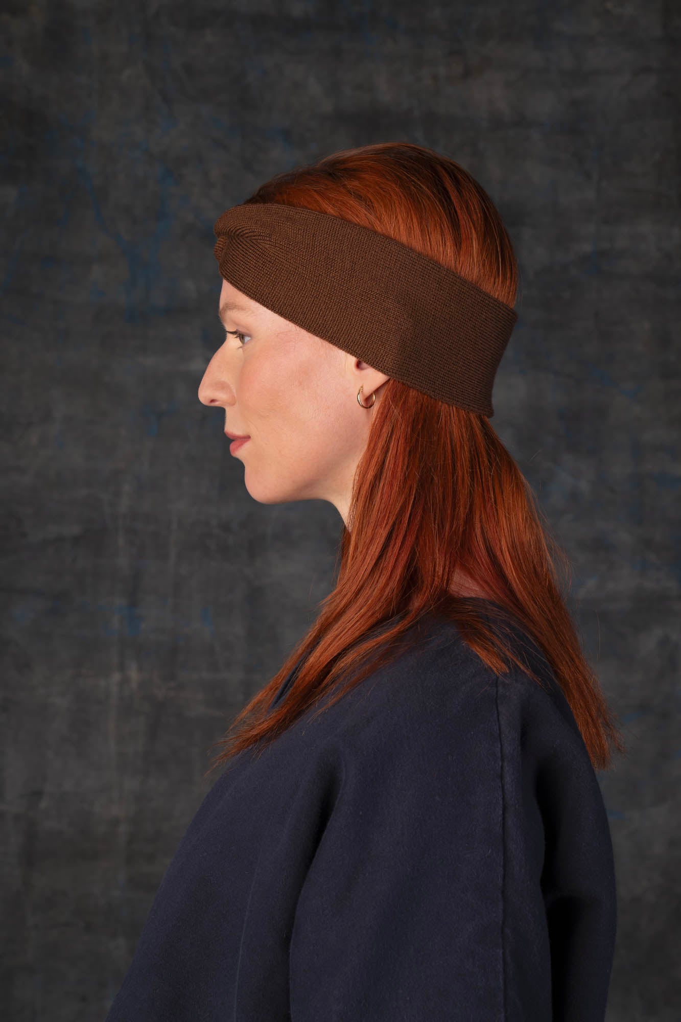 Person wearing a cacao brown knit headband and dark blue coat against a dark background.