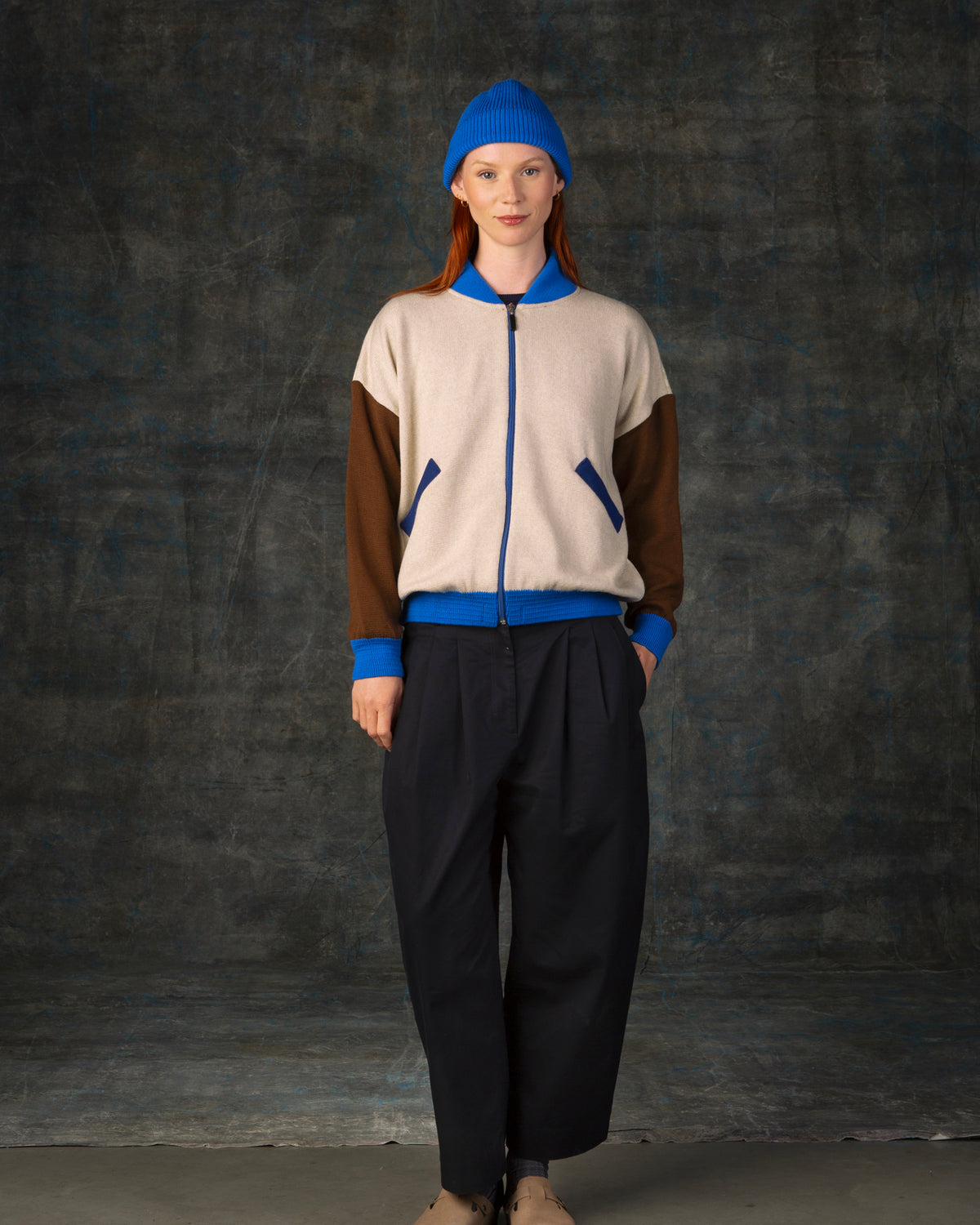 Female model wearing an ecru and brown knit bomber jacket with cobalt blue details and matching cobalt blue beanie.