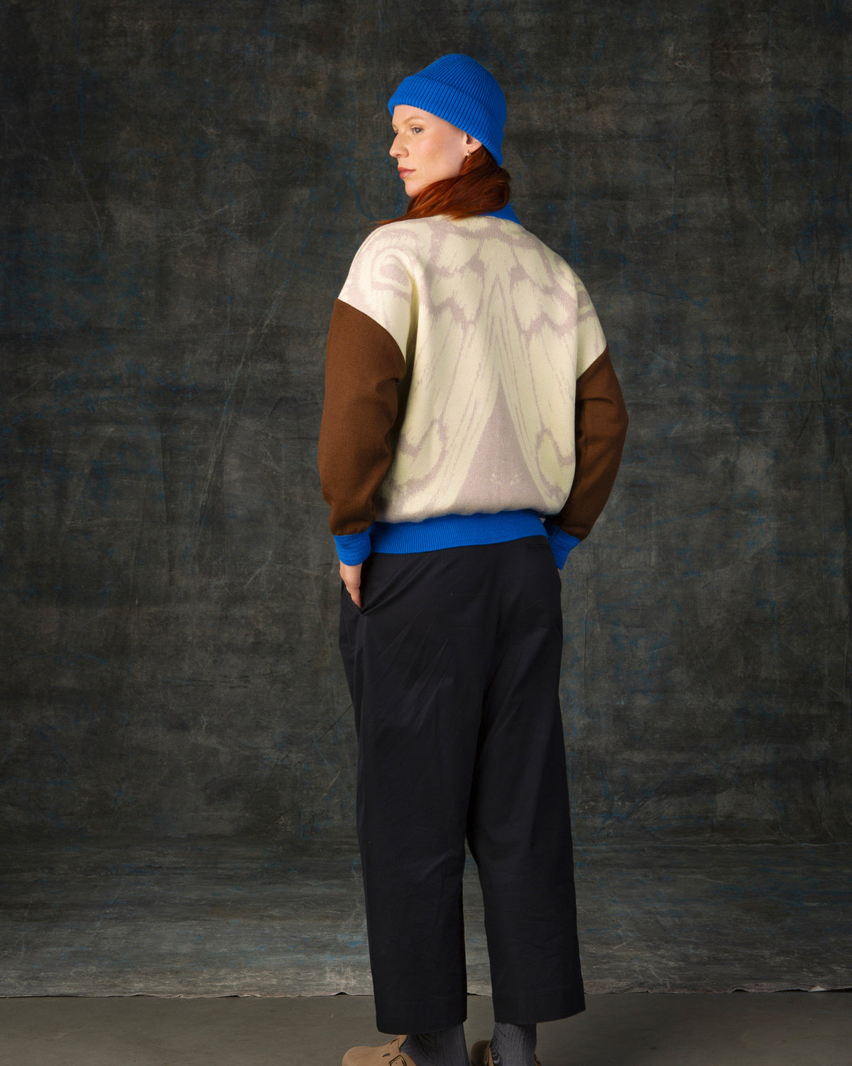 Female model facing away from the camera wearing an ecru and brown bomber jacket with cobalt blue details and matching cobalt blue beanie. There is a subtle organic pattern inspired by a butterfly's wings on the back of the jacket.