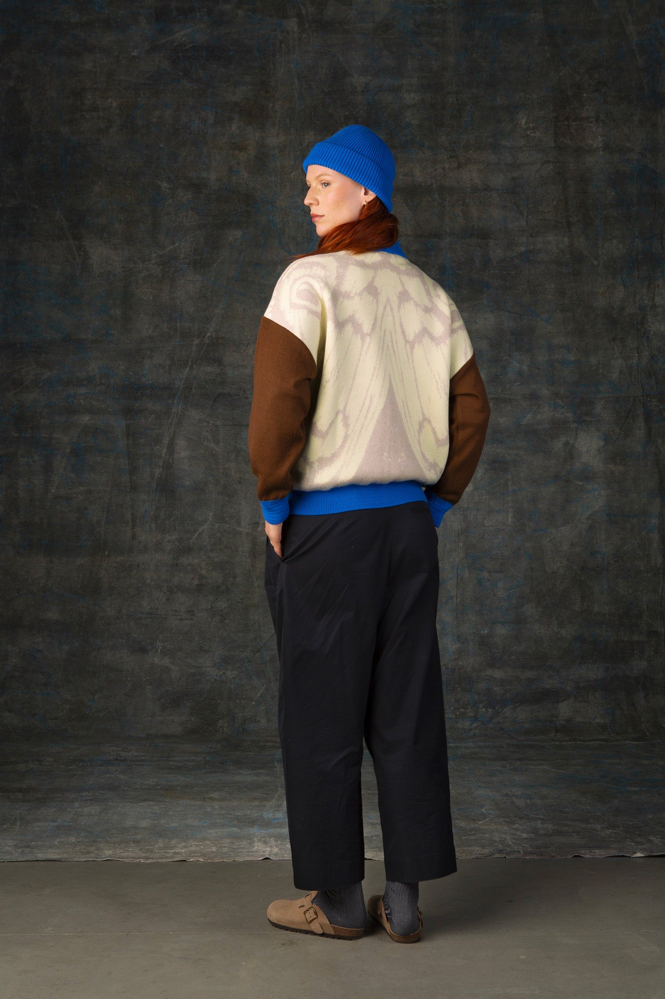 Female model facing away from the camera wearing an ecru and brown bomber jacket with cobalt blue details and matching cobalt blue beanie. There is a subtle organic pattern inspired by a butterfly's wings on the back of the jacket.