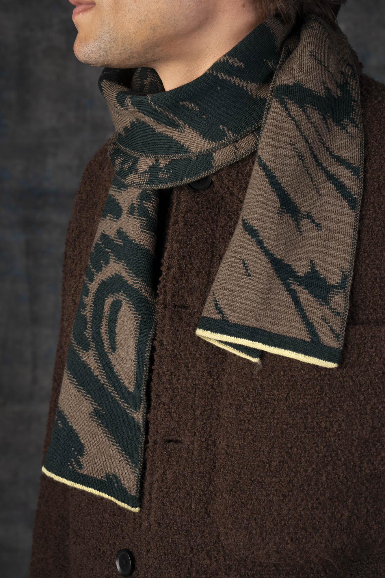 Close-up of a male model wearing a brown jacket with a green and olive maxi scarf, with an organic pattern, draped around his neck.