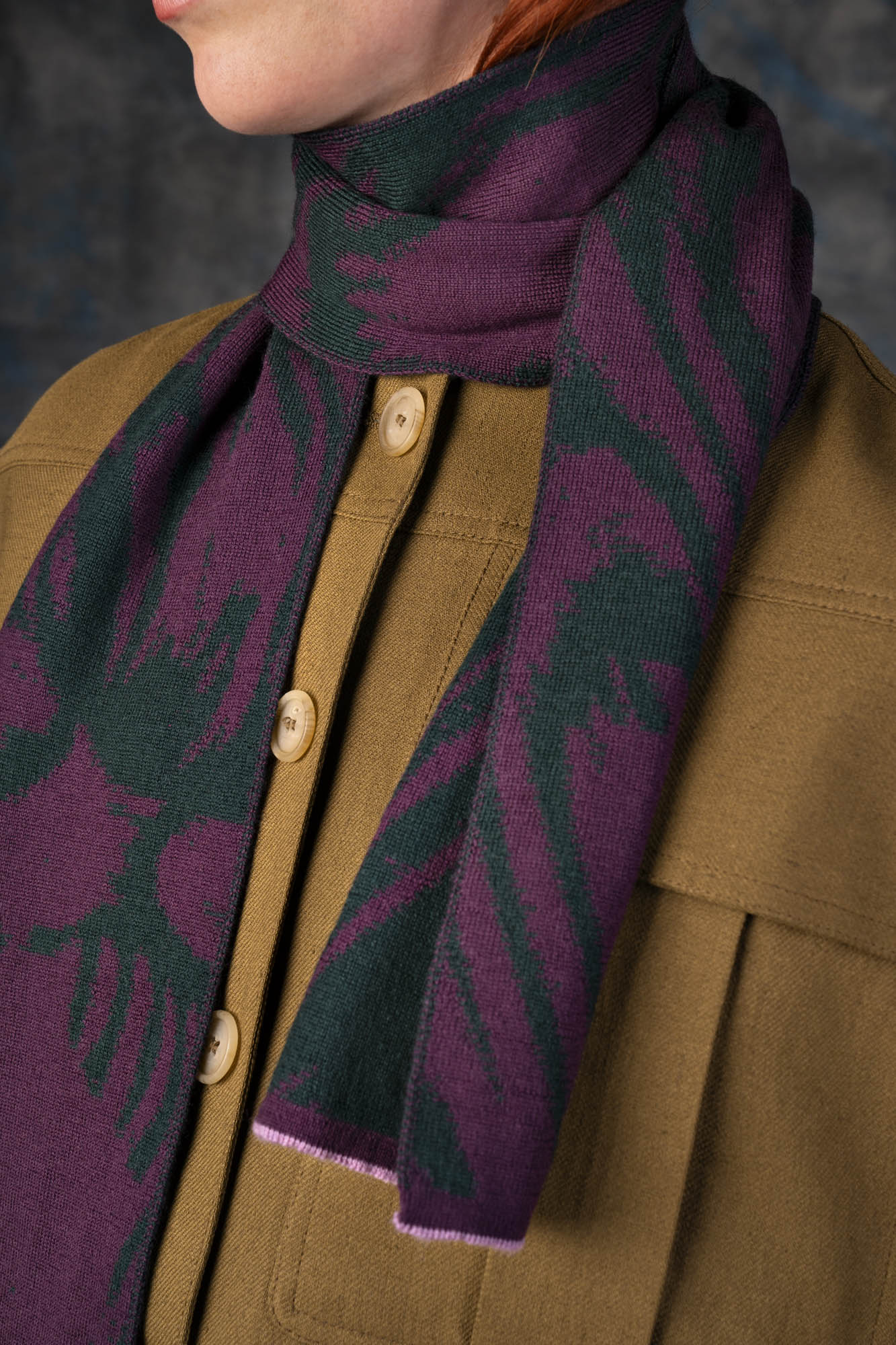 Close-up of a female model wearing an olive green jacket with a purple and forest green mini scarf, with nature-inspired pattern, draped around her neck.