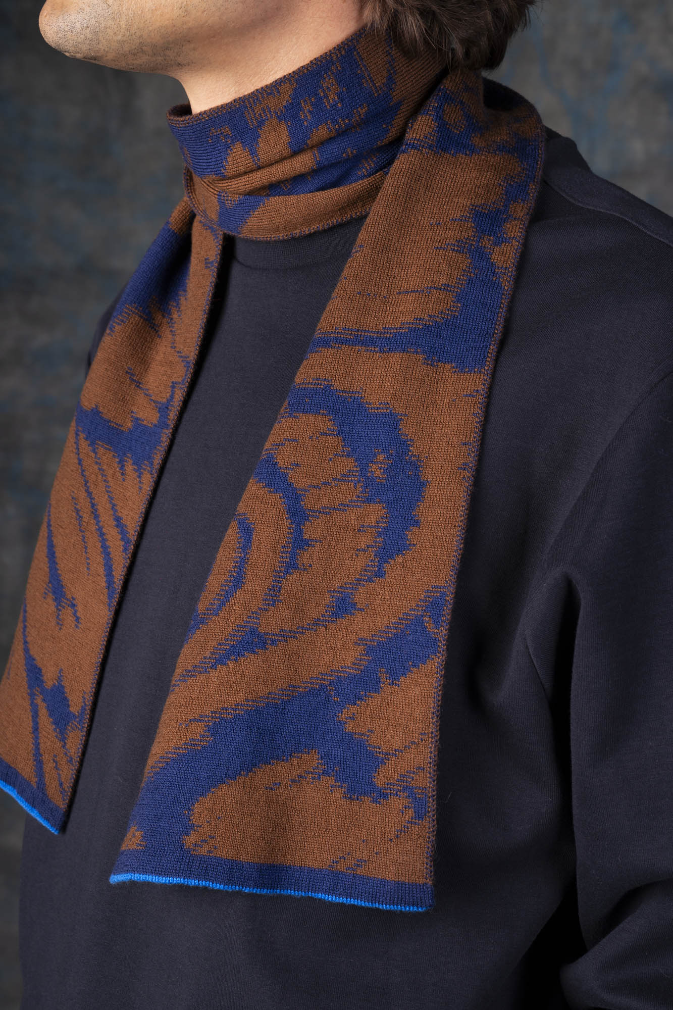 Close-up of a male model wearing a dark blue jumper with a brown and blue mini scarf, with an organic pattern, draped around his neck.