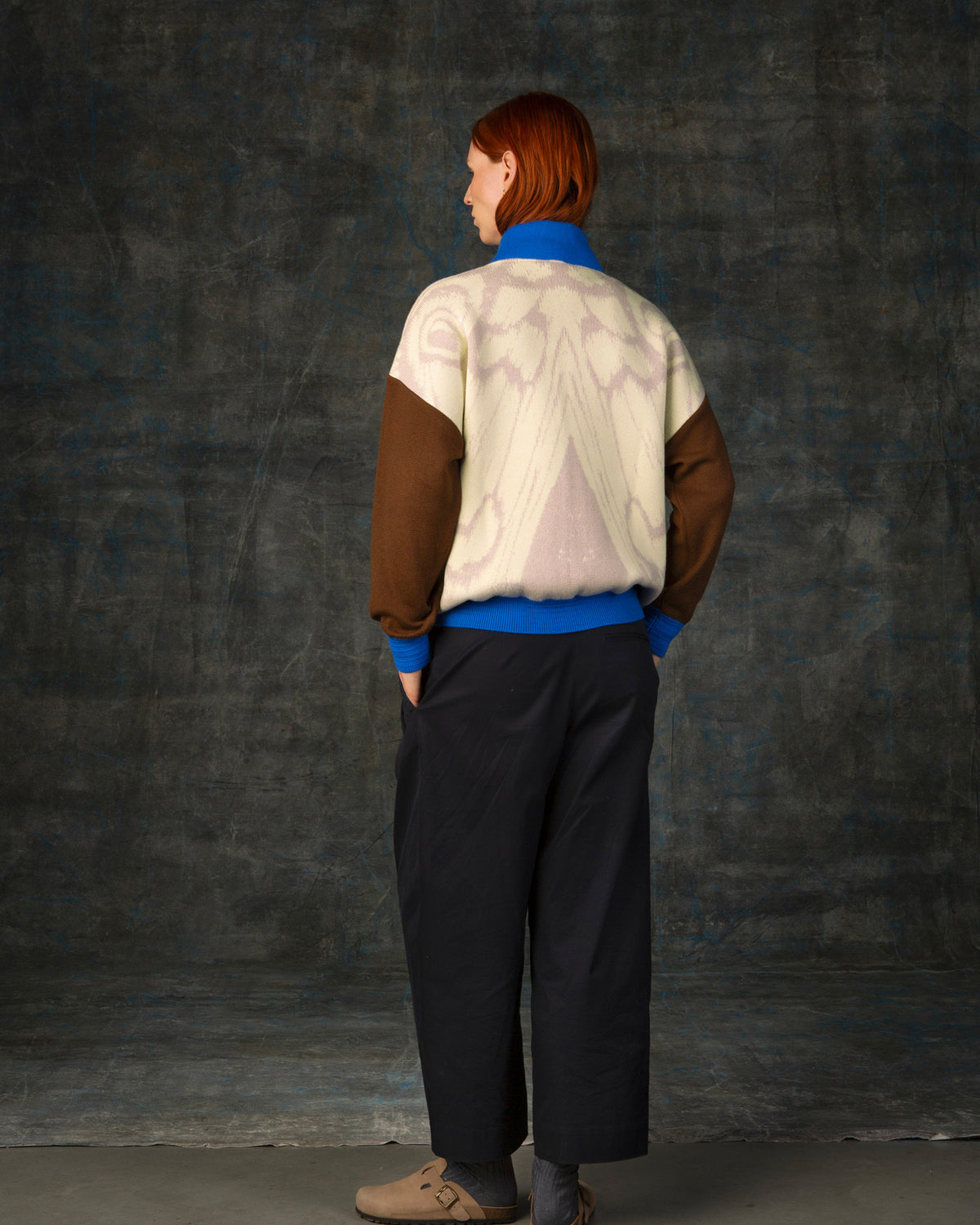 Female model facing away from the camera wearing an ecru and brown bomber jacket with cobalt blue details. There is a subtle organic pattern inspired by a butterfly's wings on the back of the jacket.