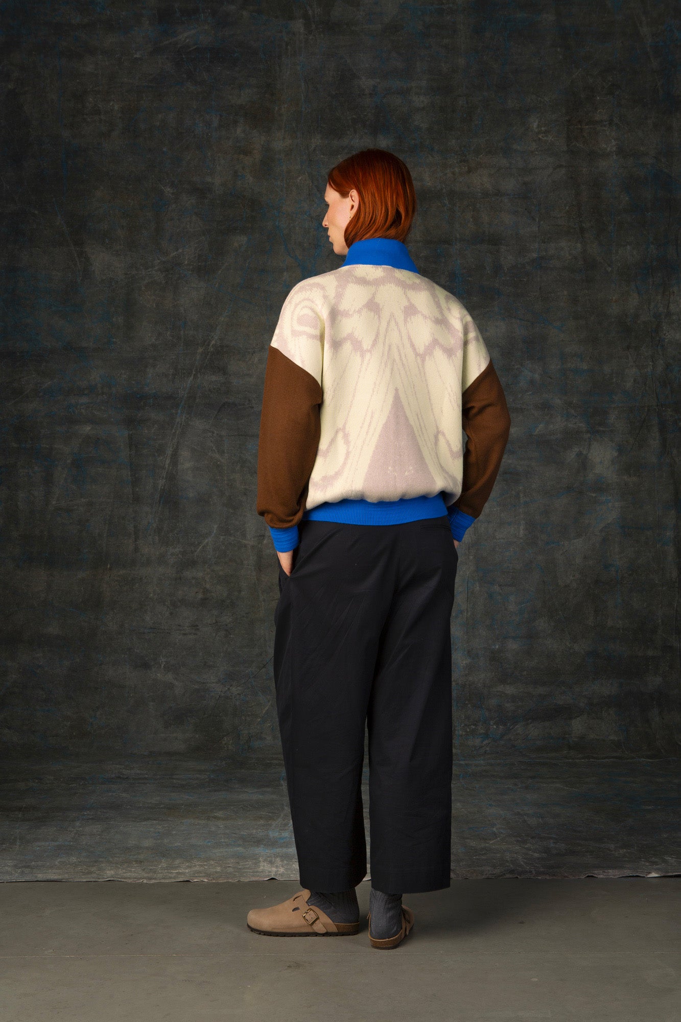 Female model facing away from the camera wearing an ecru and brown bomber jacket with cobalt blue details. There is a subtle organic pattern inspired by a butterfly's wings on the back of the jacket.