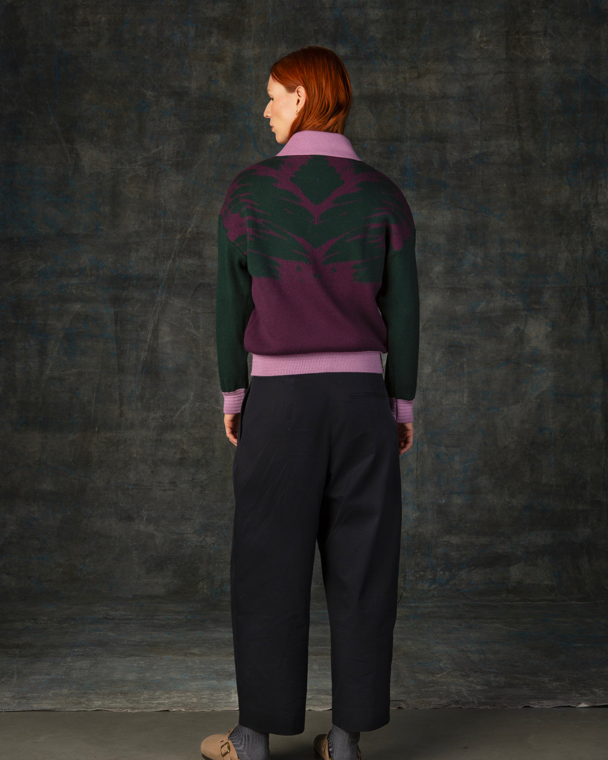 Female model wearing a grape purple bomber jacket with dark green sleeves and pink cuffs. There is an organic pattern inspired by a butterfly's wings on the back of the jacket.