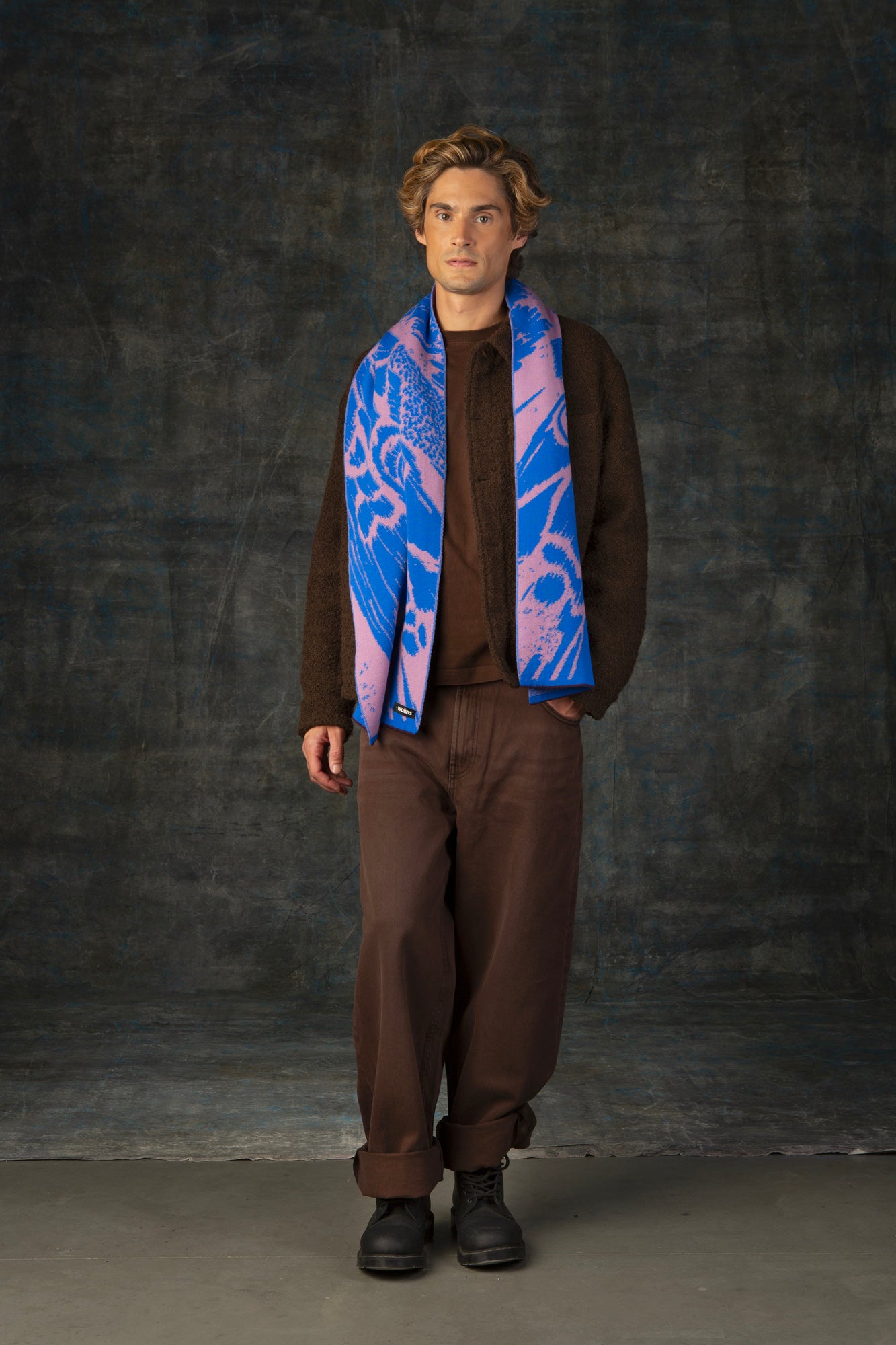 Male model wearing a brown outfit with a cobalt blue and pink midi scarf draped over his shoulders, against a dark background.