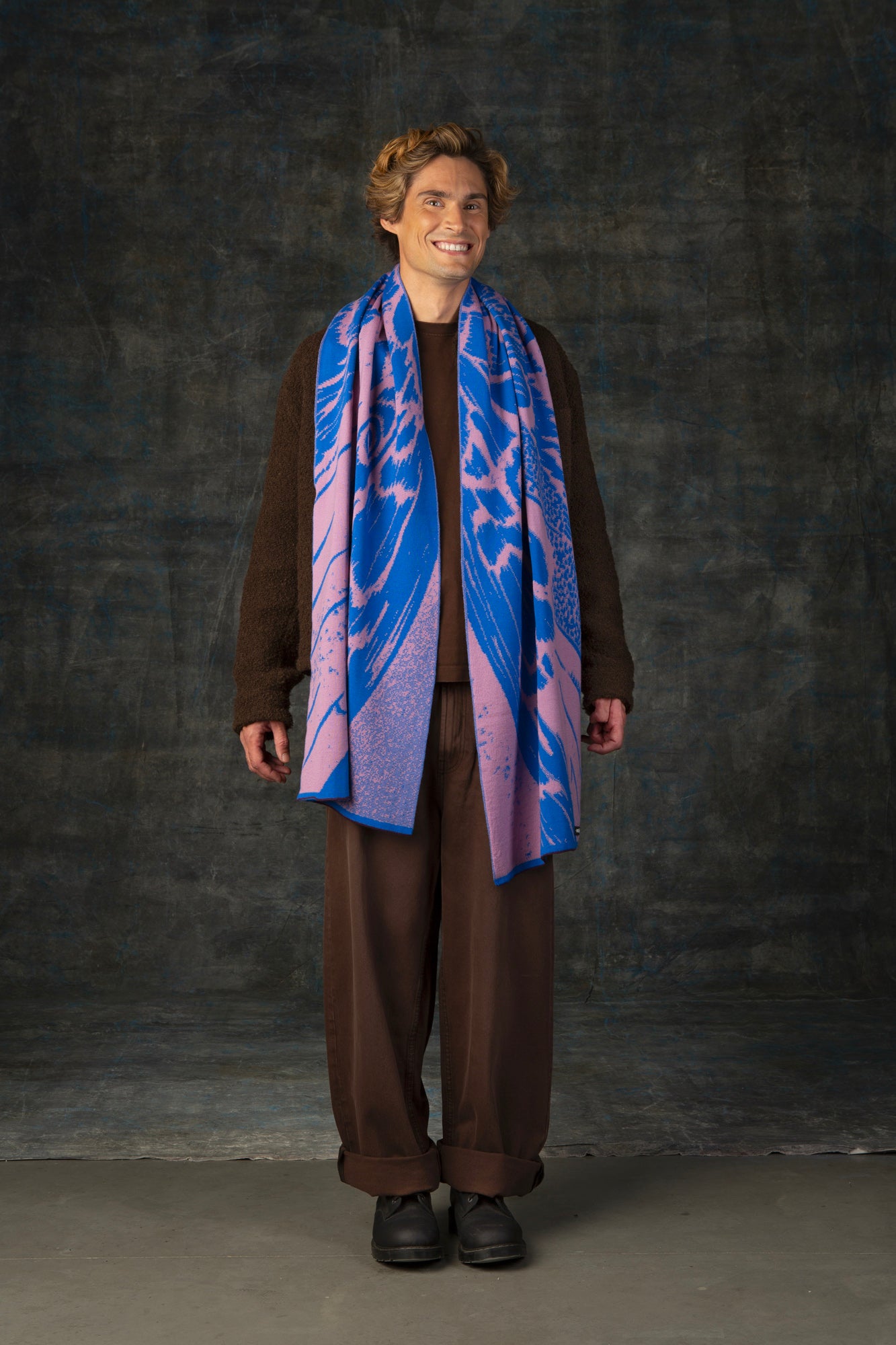 Male model wearing a brown outfit with a cobalt blue and pink maxi scarf draped over his shoulders, against a dark background.