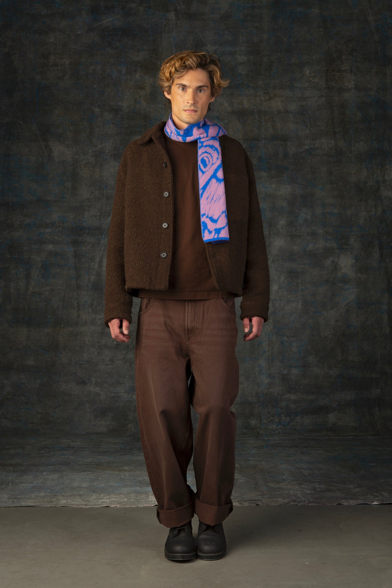 Male model wearing a brown outfit with a cobalt blue and pink mini scarf draped around his neck, against a dark background.