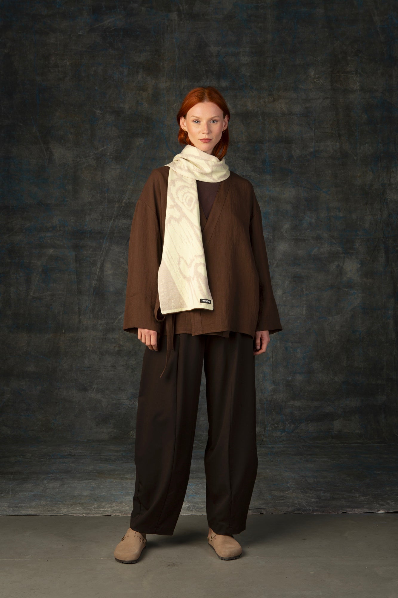 Female model wearing a brown outfit with an ecru midi scarf draped over her shoulder, against a dark background.