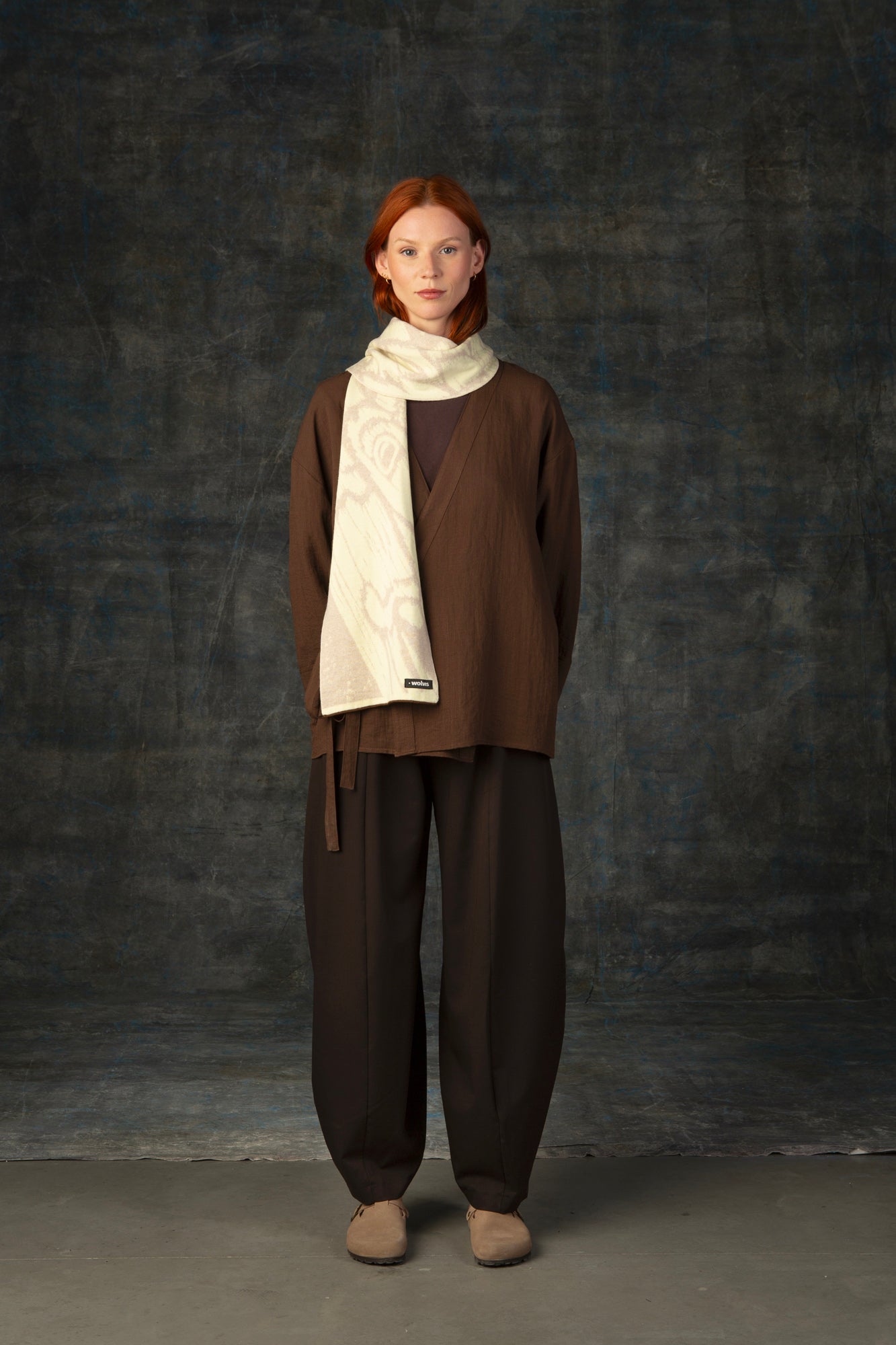 Female model wearing a brown outfit with an ecru midi scarf draped over her shoulder, against a dark background.