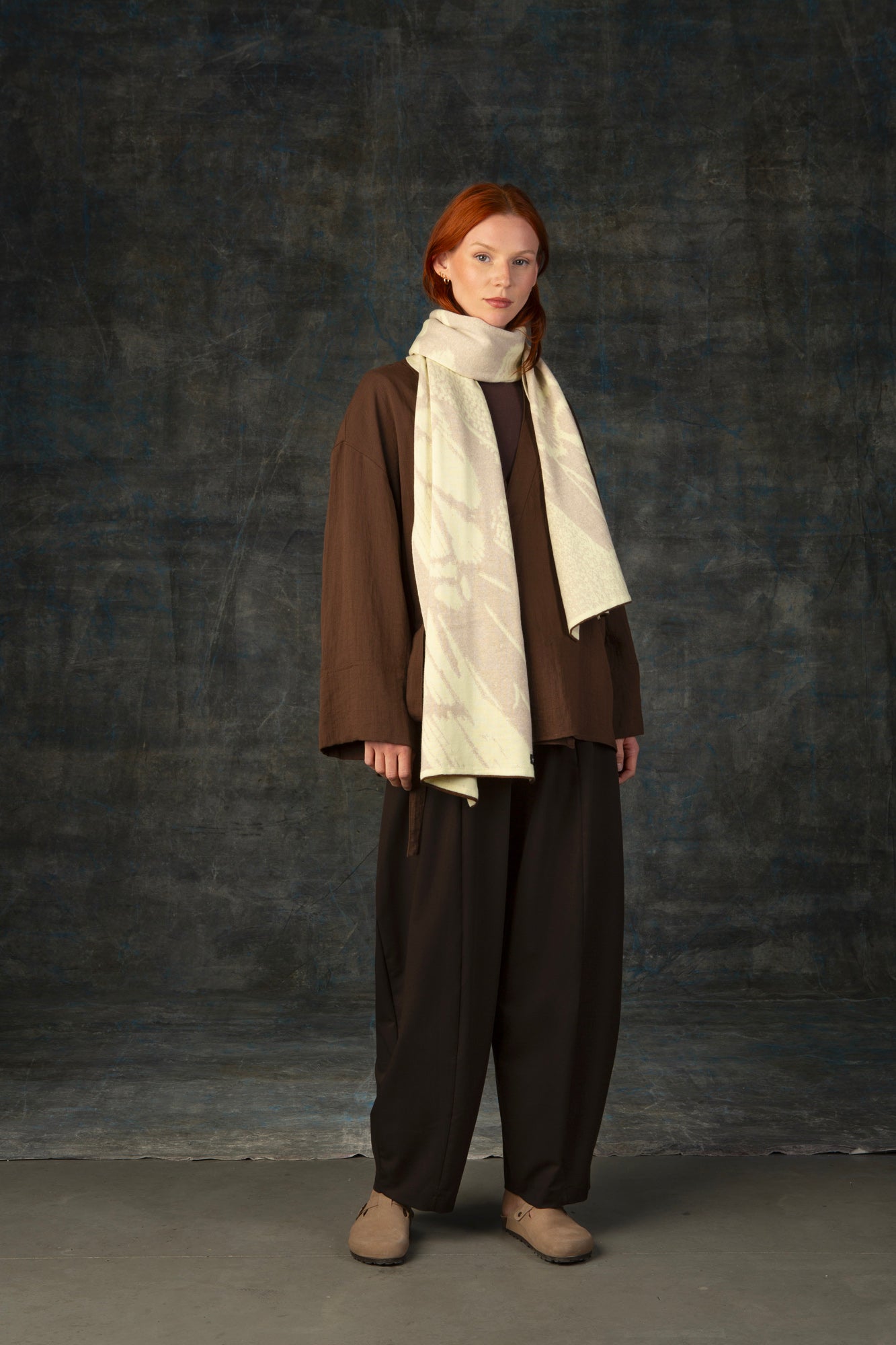 Female model wearing a brown outfit with an ecru maxi scarf draped around her neck, against a dark background.