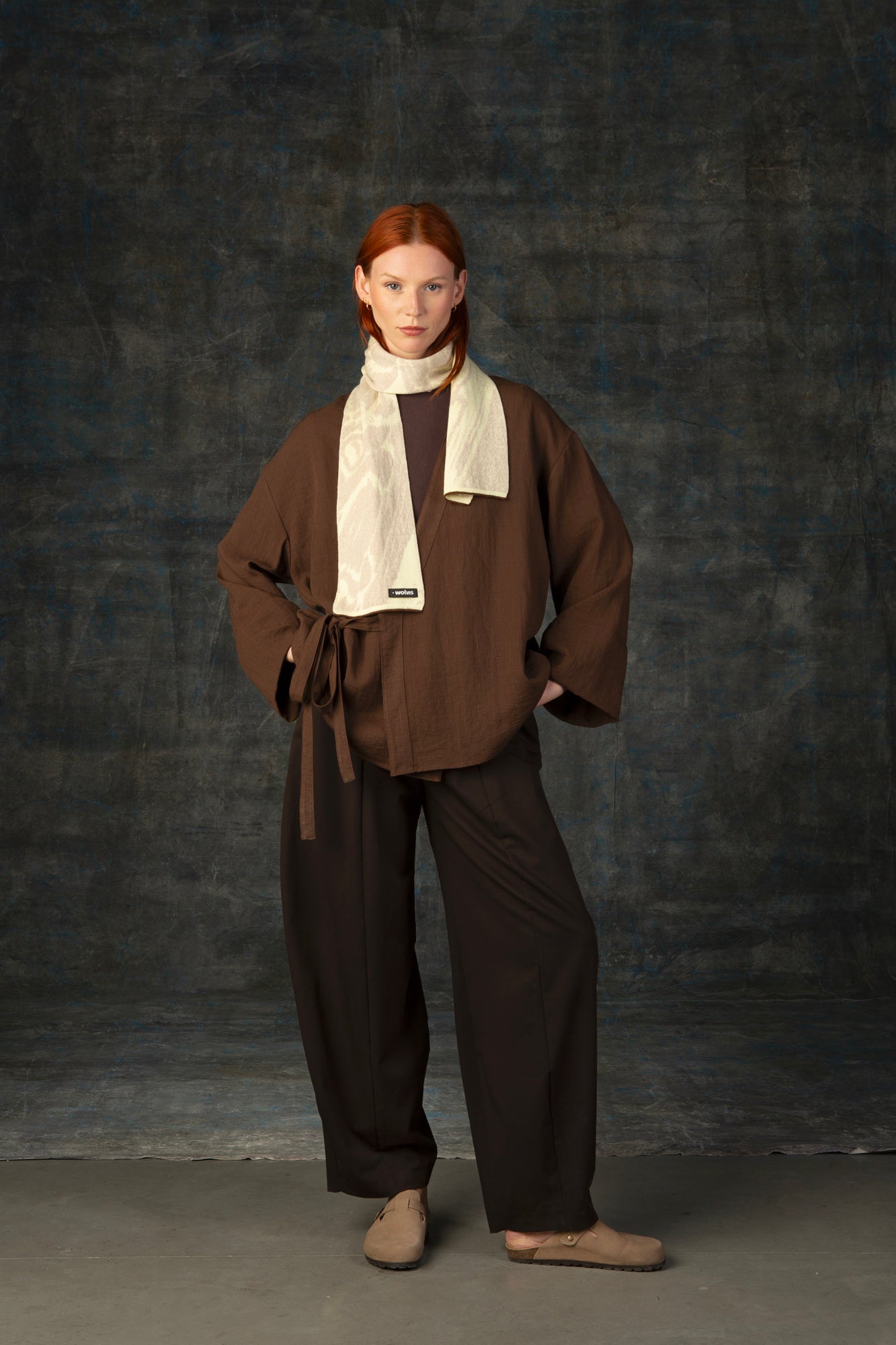 Female model wearing a brown outfit with an ecru mini scarf draped around her neck, against a dark background.