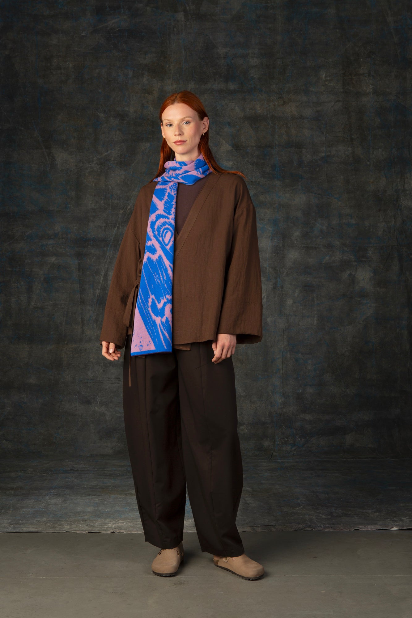Female model wearing a brown outfit with a cobalt blue and pink midi scarf draped around her neck, against a dark background.