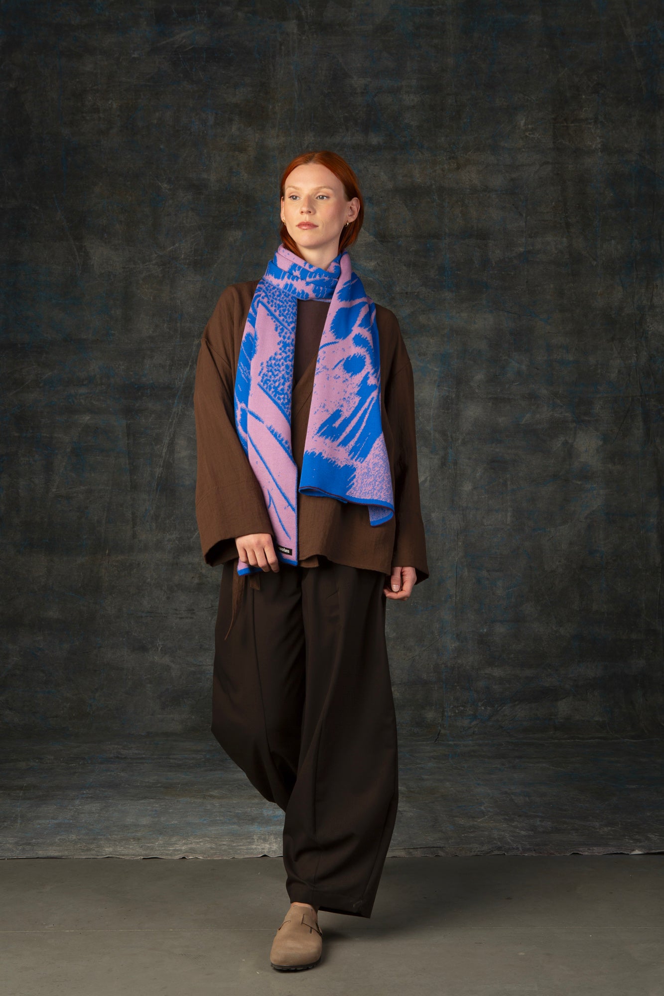 Female model wearing a brown outfit with a cobalt blue and pink maxi scarf draped around her neck, against a dark background.