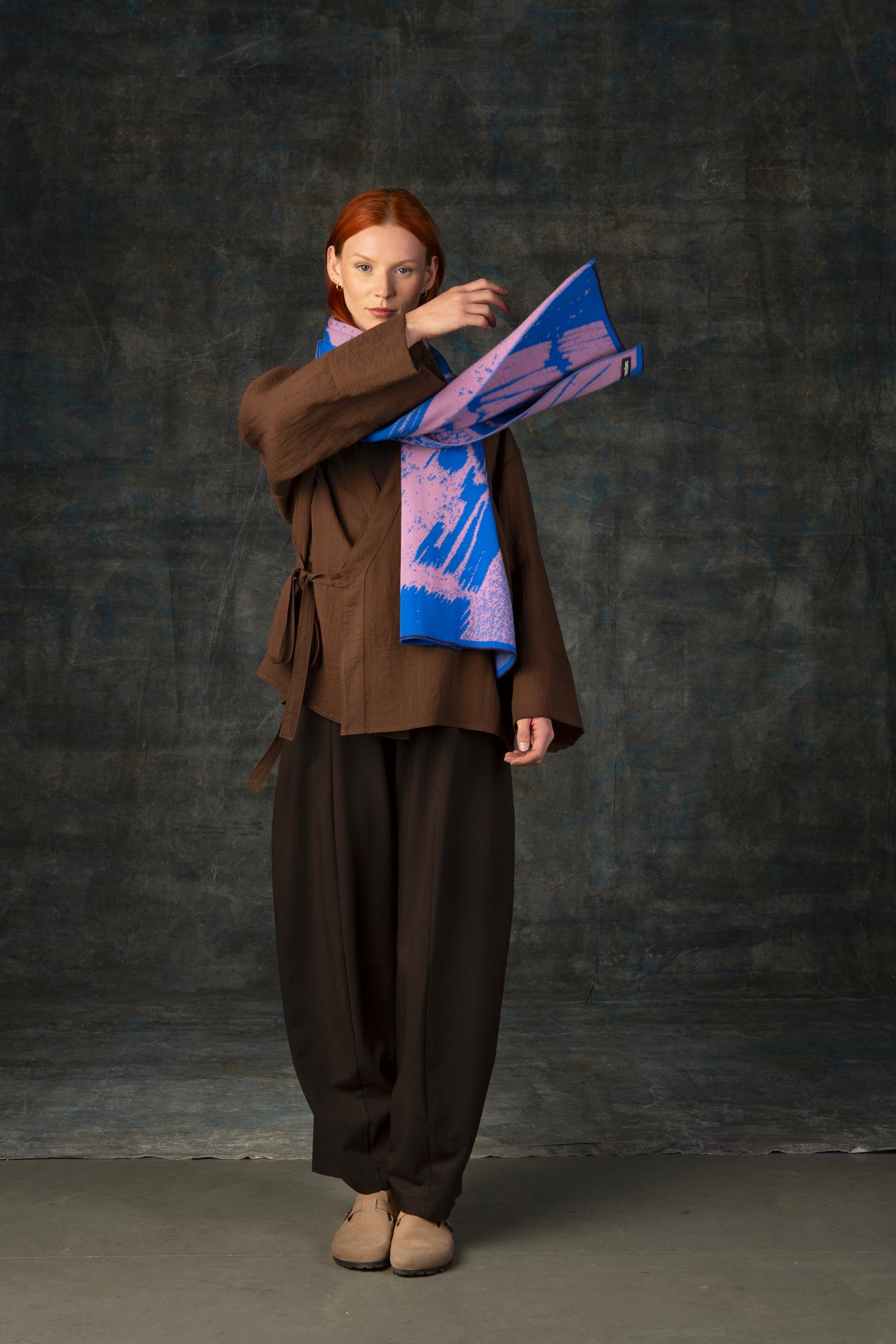 Female model wearing a brown outfit throwing on end of a cobalt blue and pink maxi scarf over her shoulder, against a dark background.
