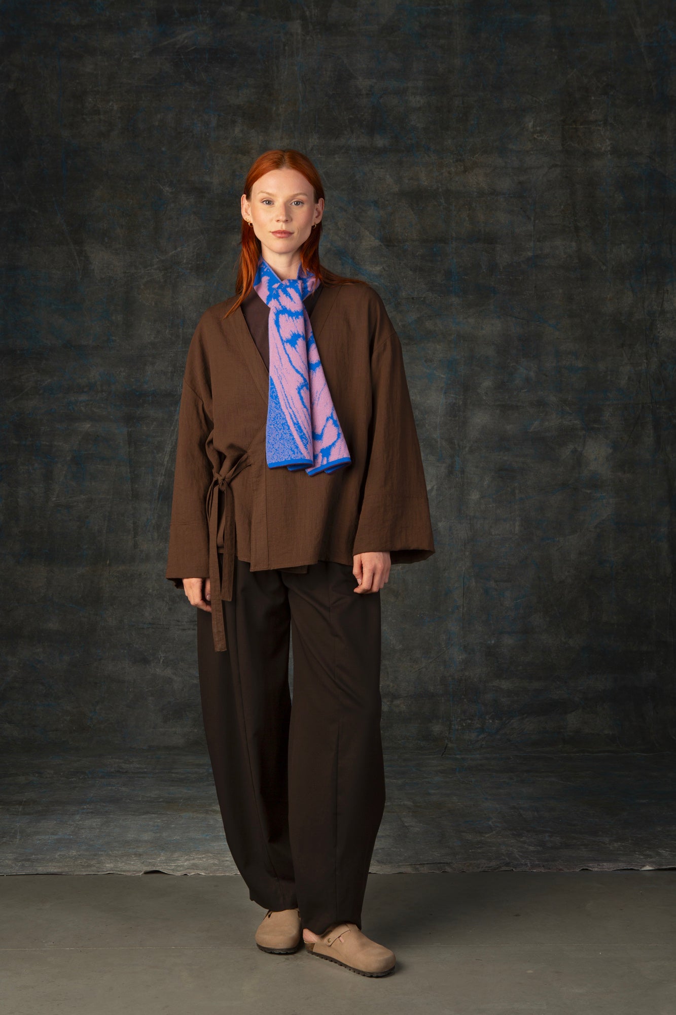 Female model wearing a brown outfit with a cobalt blue and pink mini scarf loosely tied around her neck, against a dark background.