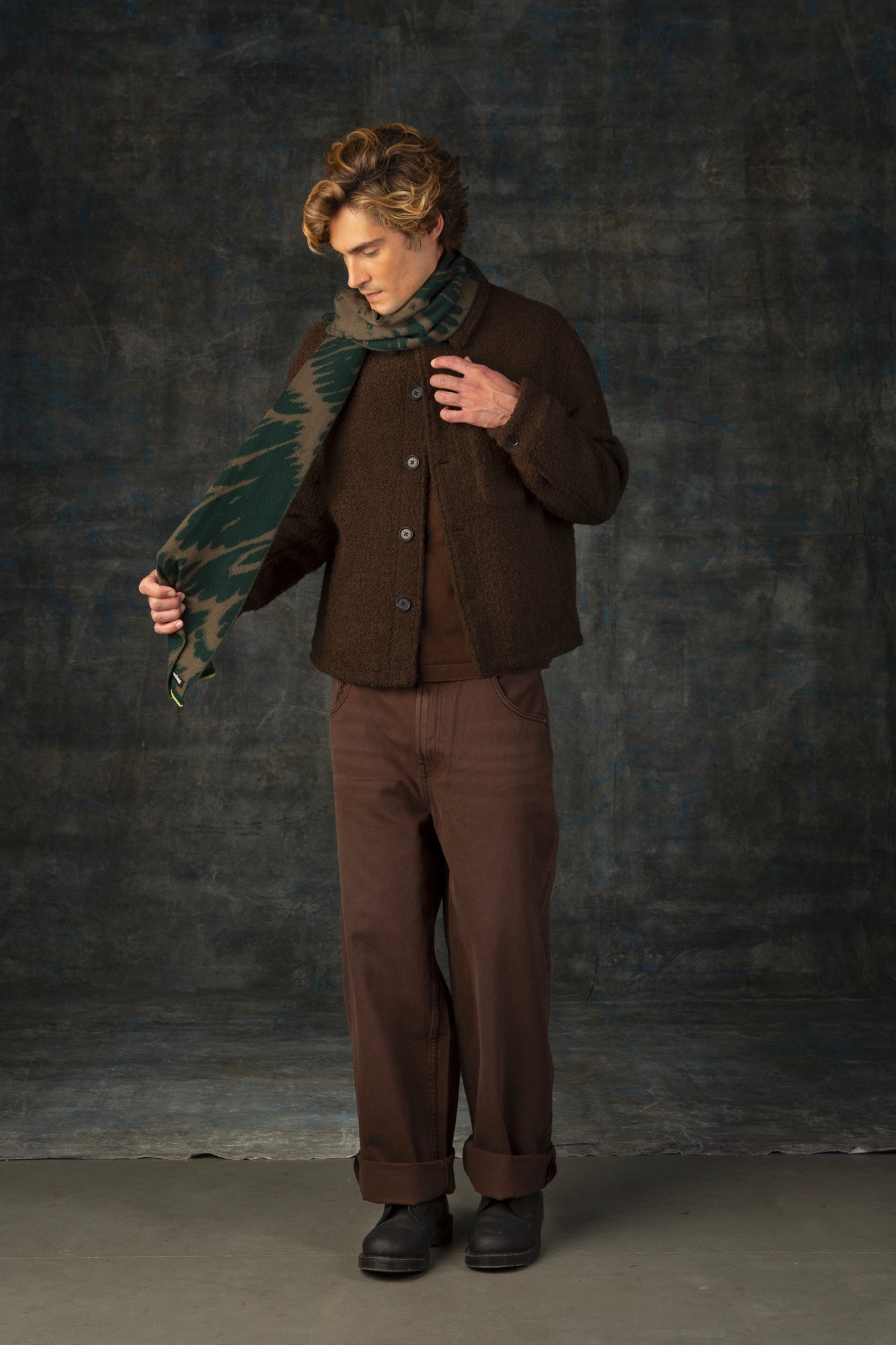 Male model wearing a brown outfit with a green and olive maxi scarf, with an organic pattern, draped around his neck.