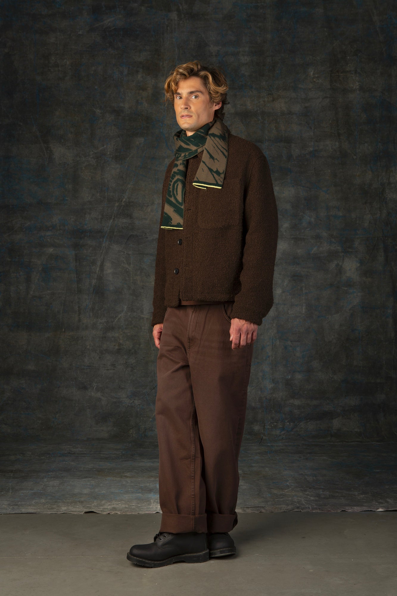 Male model wearing a brown outfit with a green and olive mini scarf, with an organic pattern, draped around his neck.
