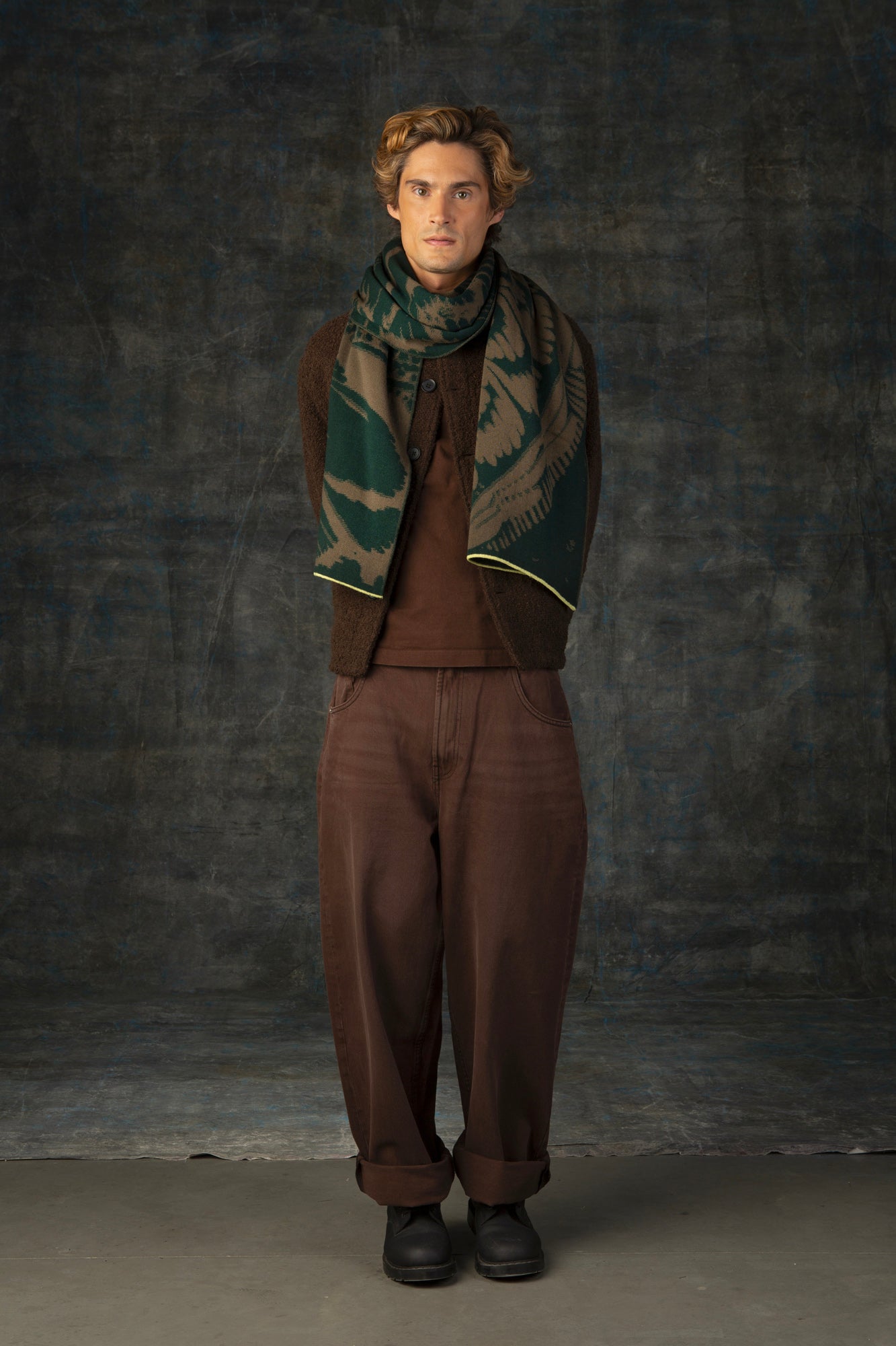 Male model wearing a brown outfit with a green and olive maxi scarf, with an organic pattern, draped around his neck.