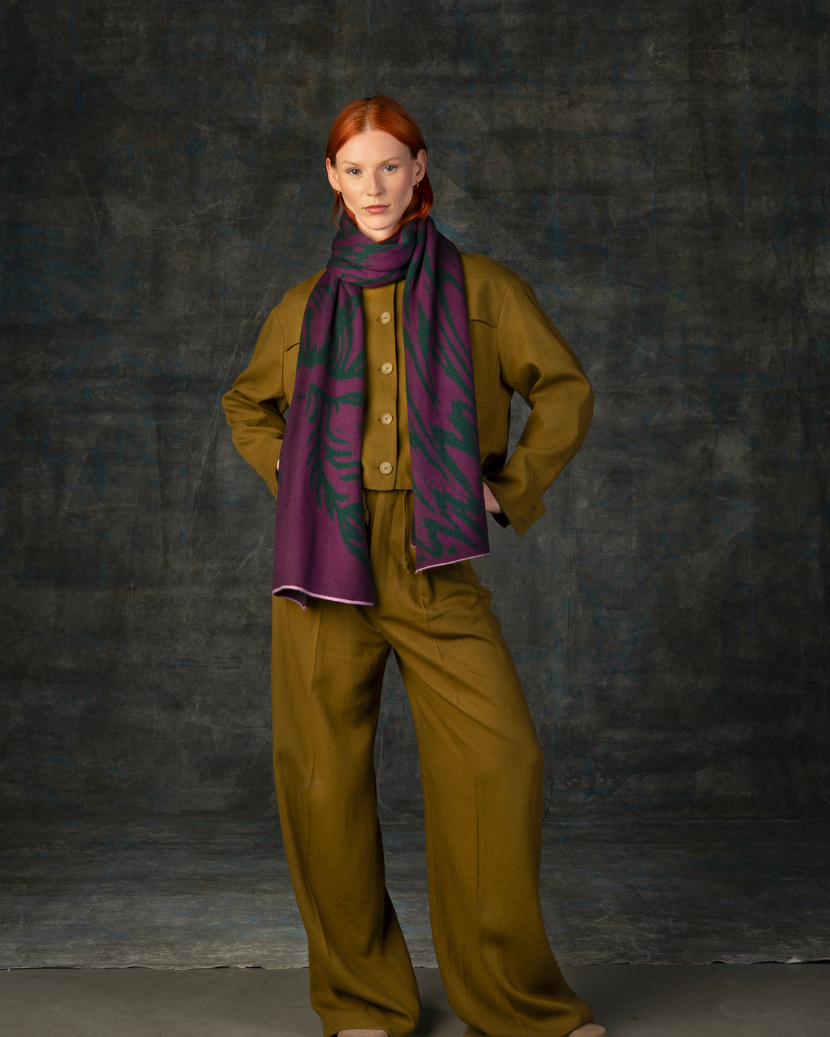 Female model wearing an olive green outfit with a purple and forest green maxi scarf, with nature-inspired pattern, draped around her neck.