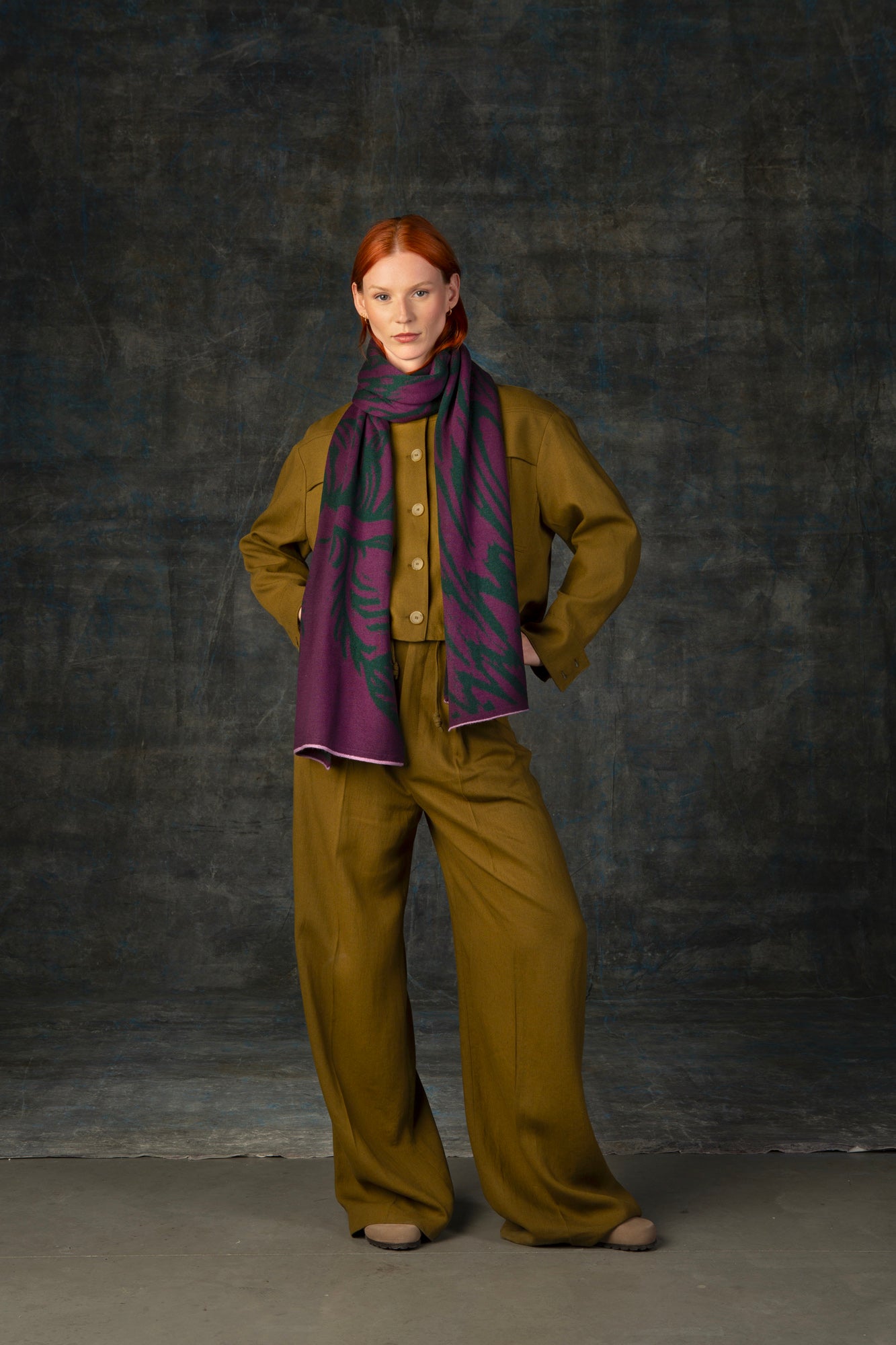 Female model wearing an olive green outfit with a purple and forest green maxi scarf, with nature-inspired pattern, draped around her neck.