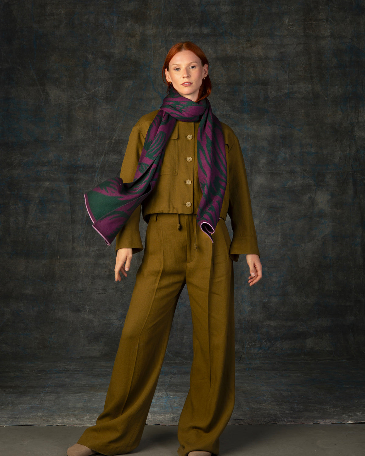 Female model wearing an olive green outfit with a purple and forest green maxi scarf, with nature-inspired pattern, draped around her neck.