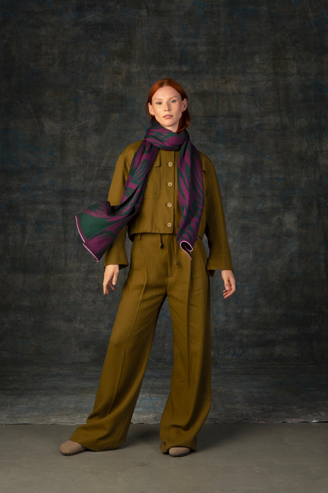 Female model wearing an olive green outfit with a purple and forest green maxi scarf, with nature-inspired pattern, draped around her neck.