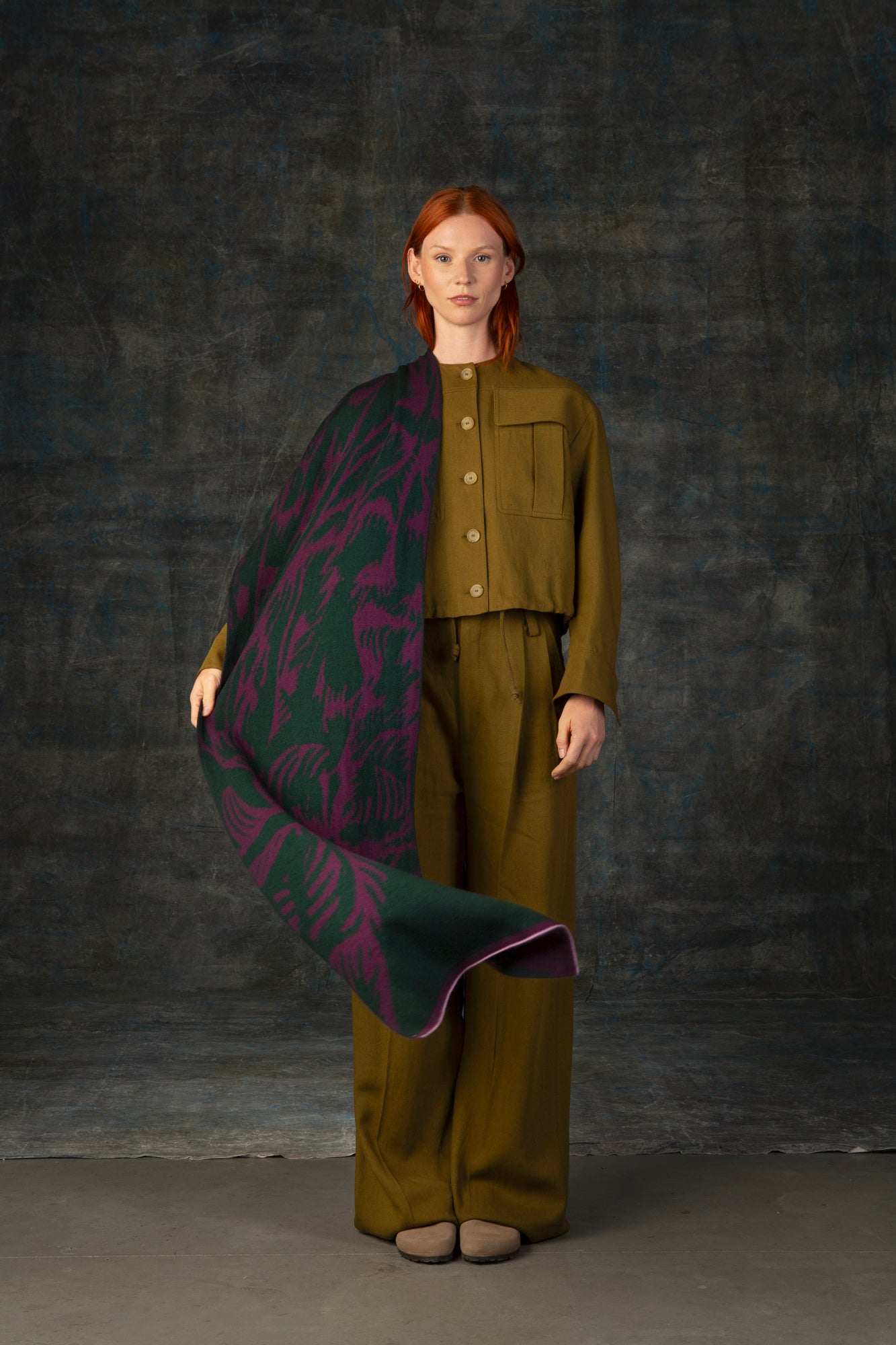 Female model wearing an olive green outfit waving a purple and forest green maxi scarf, with nature-inspired pattern, draped over her shoulder.
